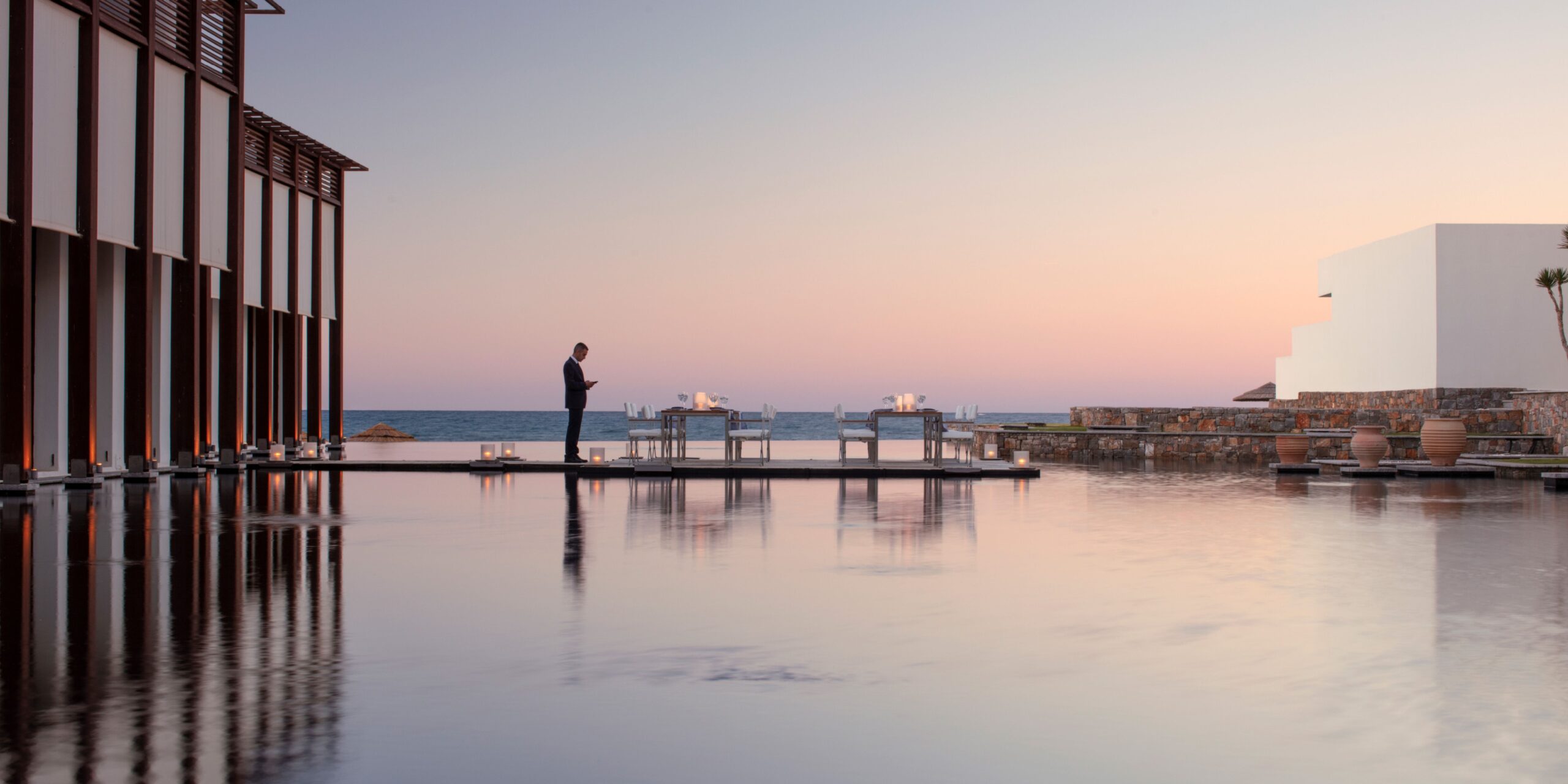 Evening scene at a resort with a reflective pool, dining tables set up, and a person standing by the water's edge, with a view of the ocean and a pastel sky.