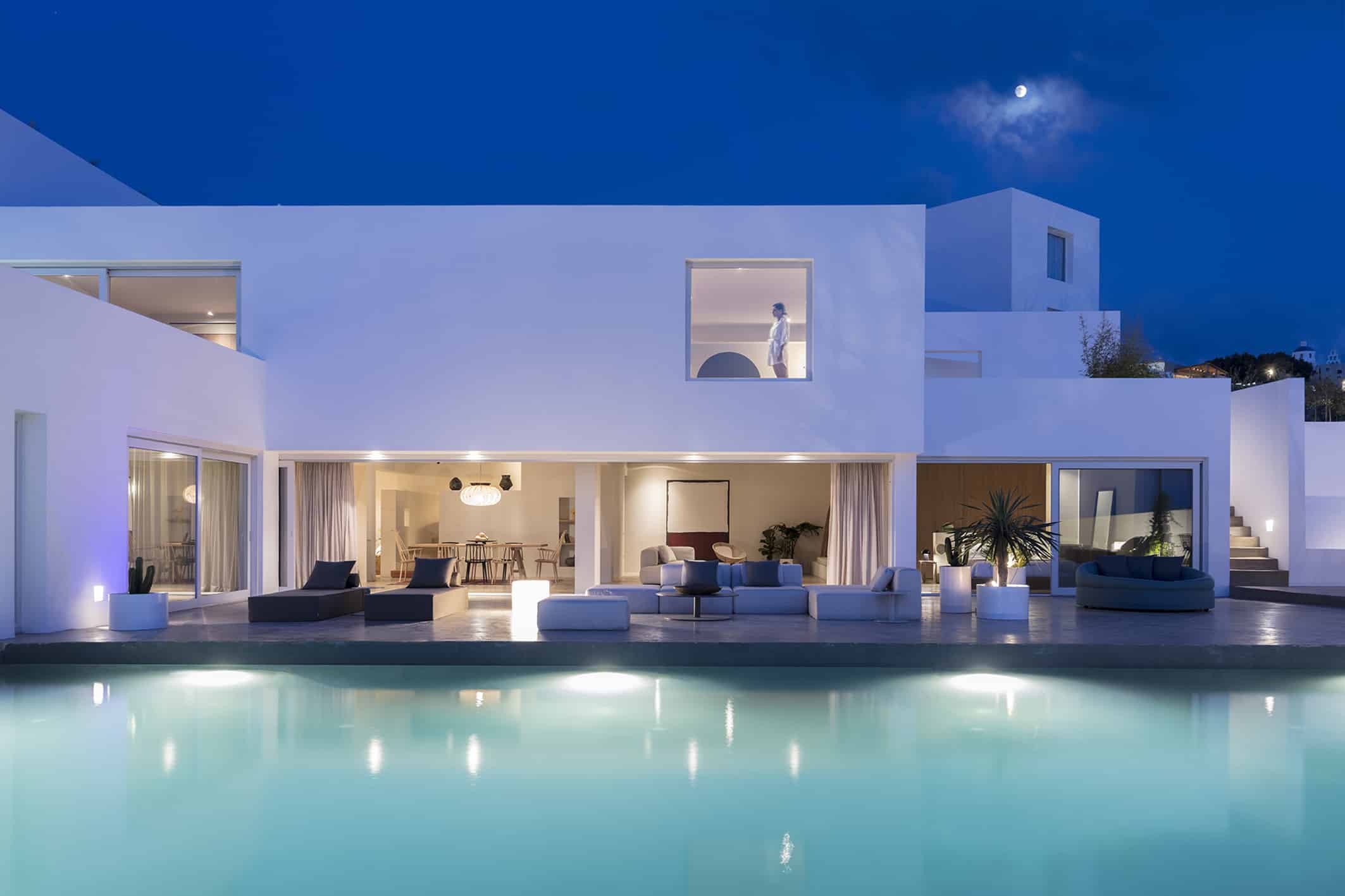 A modern villa with an open living area and pool illuminated in the evening, with a clear sky and the moon above.