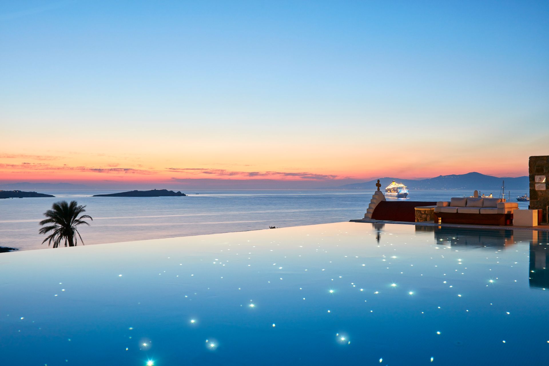 This image shows a beautiful infinity pool with a breathtaking view of the ocean. The pool is surrounded by lush greenery and palm trees, and the water is crystal clear. The sky is blue with a few white clouds, and the sun is setting in the distance. The overall impression is one of tranquility and relaxation.
