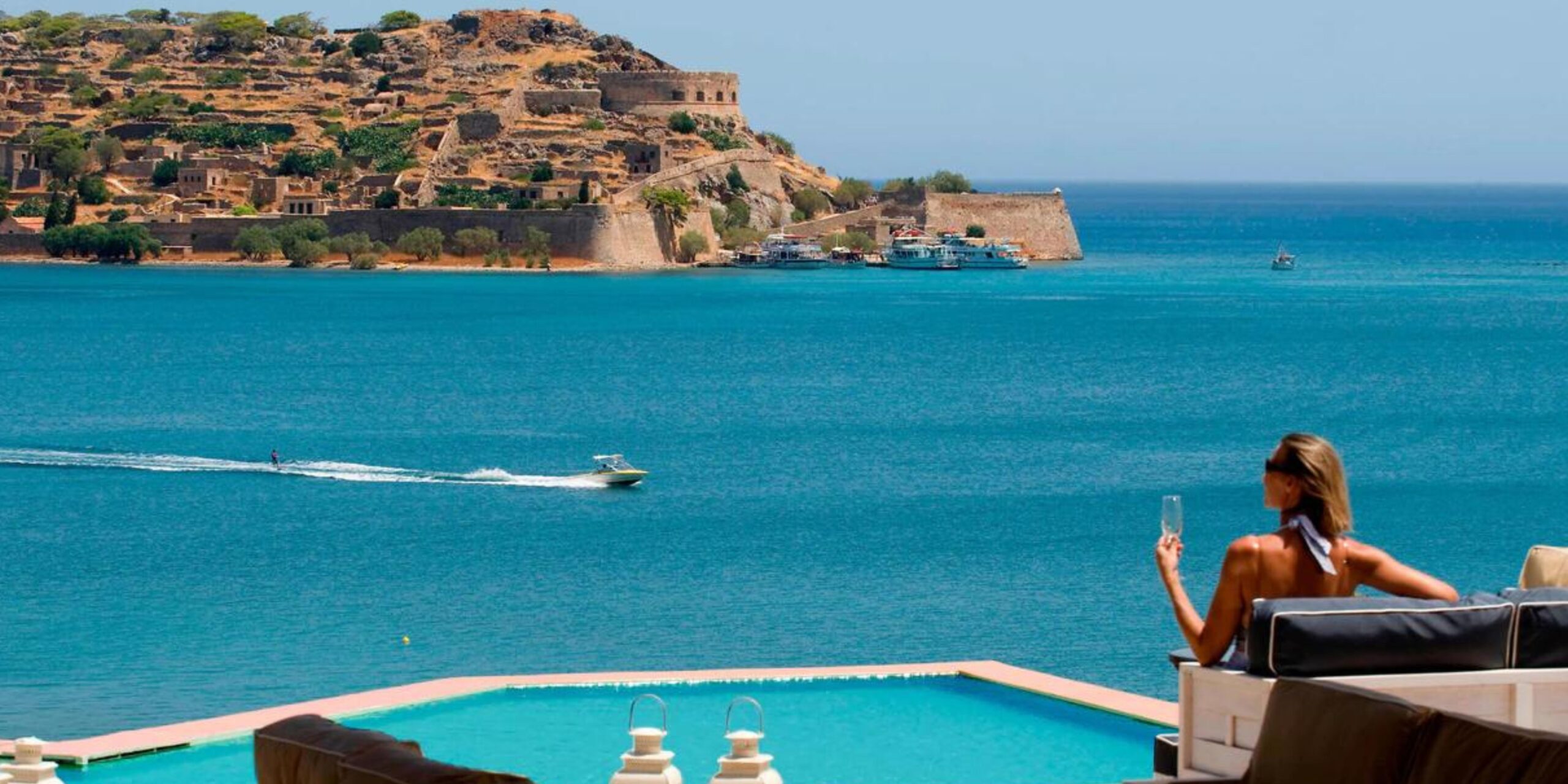 View from a poolside overlooking the sea, with a person relaxing on a lounger, speedboats in the water, and historic ruins on the hillside in the distance.