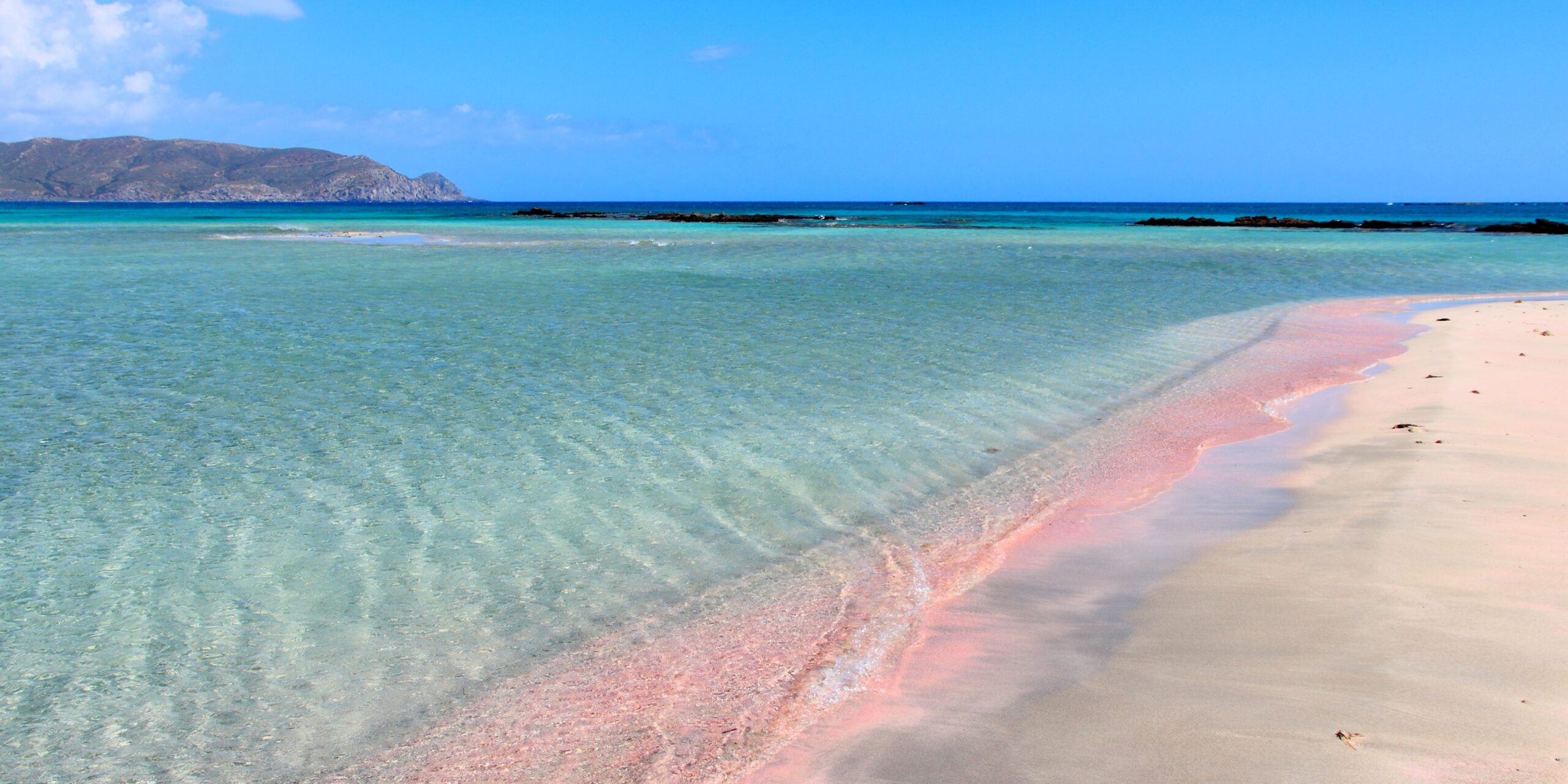 A clear turquoise sea gently laps onto a pink-tinged sandy beach with a mountainous backdrop.