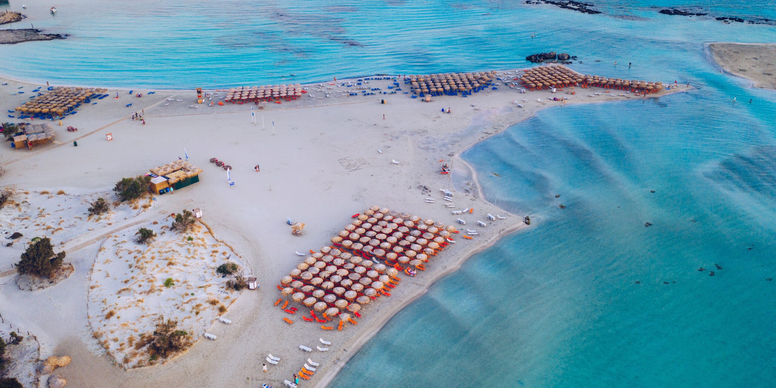 Aerial view of a sandy beach with rows of umbrellas and sun loungers, adjacent to clear blue waters.