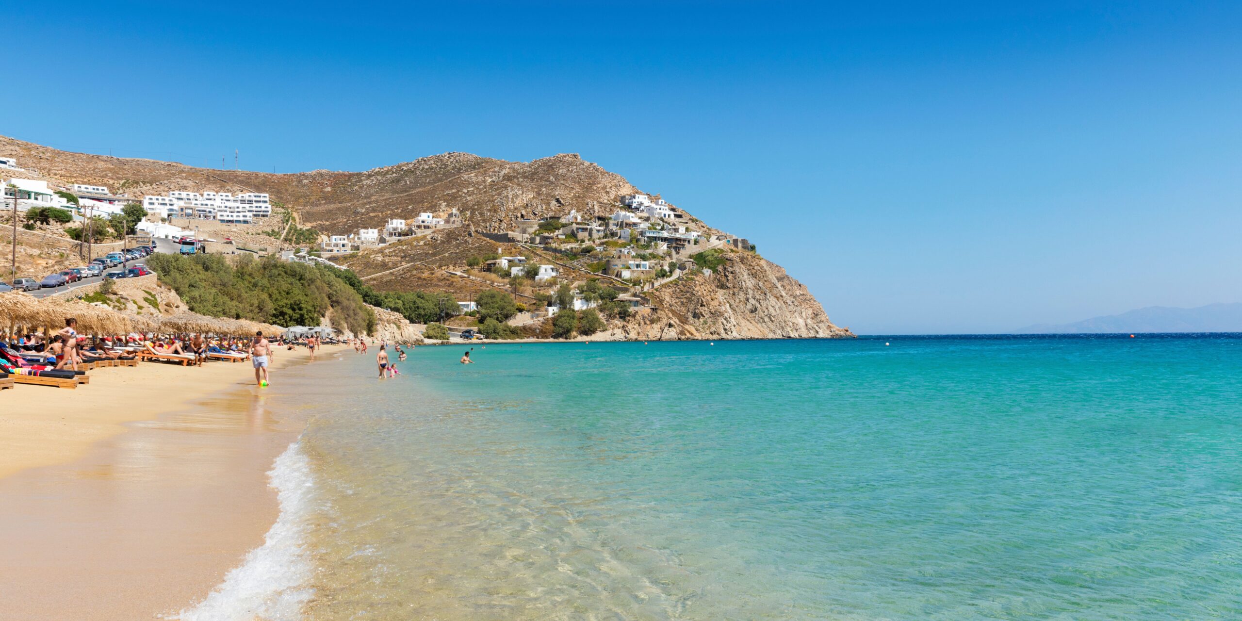 Holidaymakers bask in the sun and wade into the inviting waters of Elia Beach, framed by the scenic hills of Mykonos.