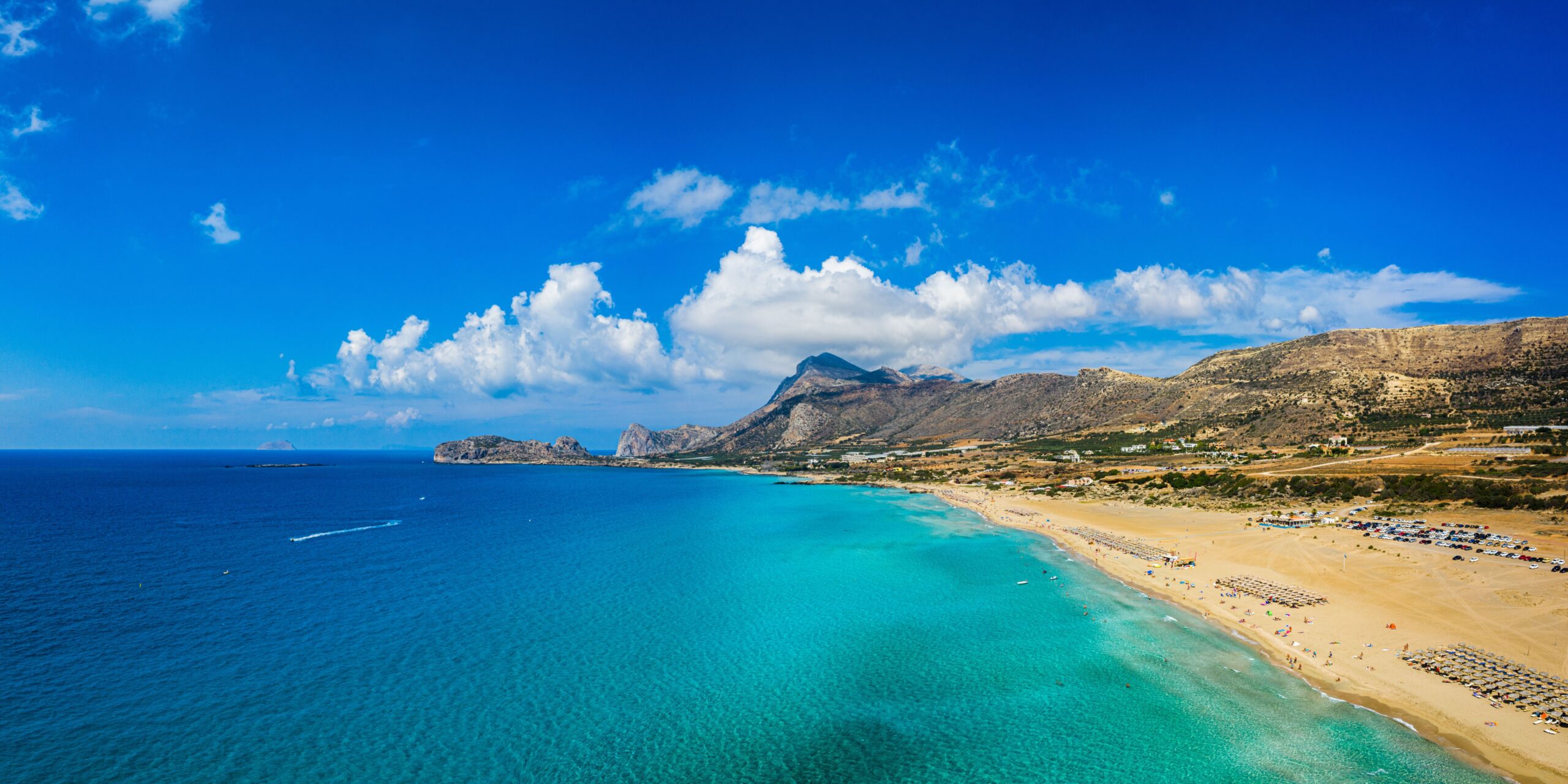 Aerial panoramic view of a long sandy beach with clear blue waters and a mountainous backdrop under a partly cloudy sky.