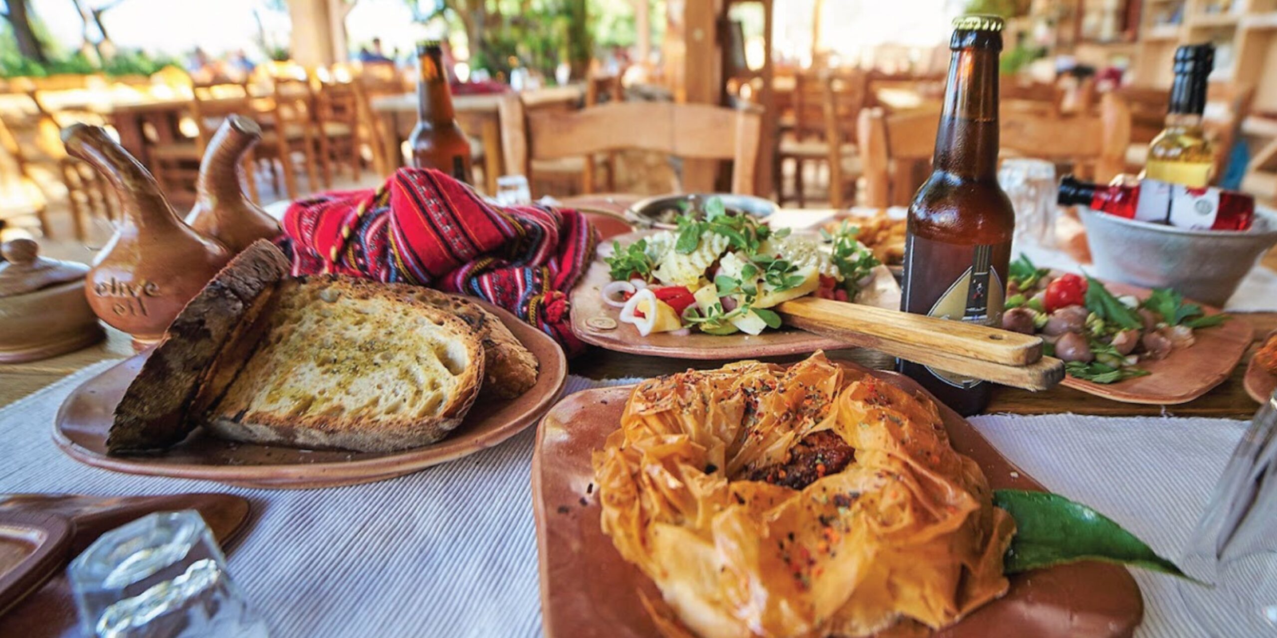 Table spread with various Greek dishes, bread, and a bottle of beer at a taverna.