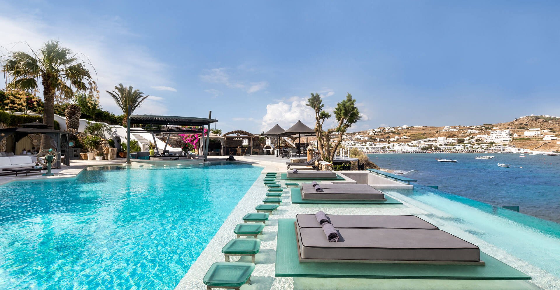 Sun-drenched Mykonos paradise: Infinity pool lined with plush sunbeds spills towards the sparkling Aegean Sea. Lush greenery and palm trees frame the picture-perfect scene.