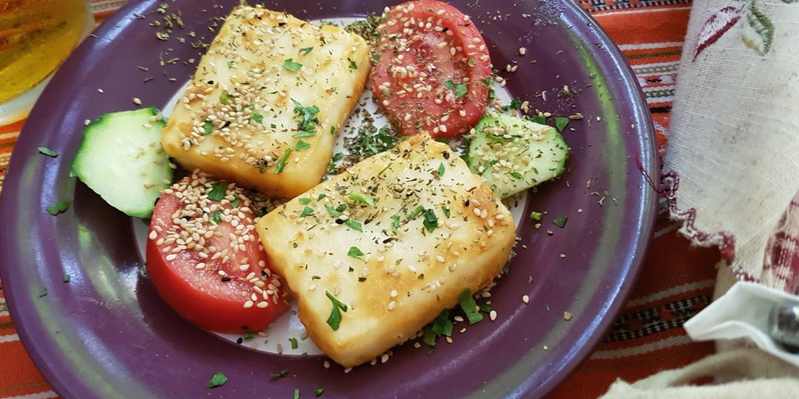 Plate with grilled cheese, tomato slices, and cucumber garnished with herbs and sesame seeds.