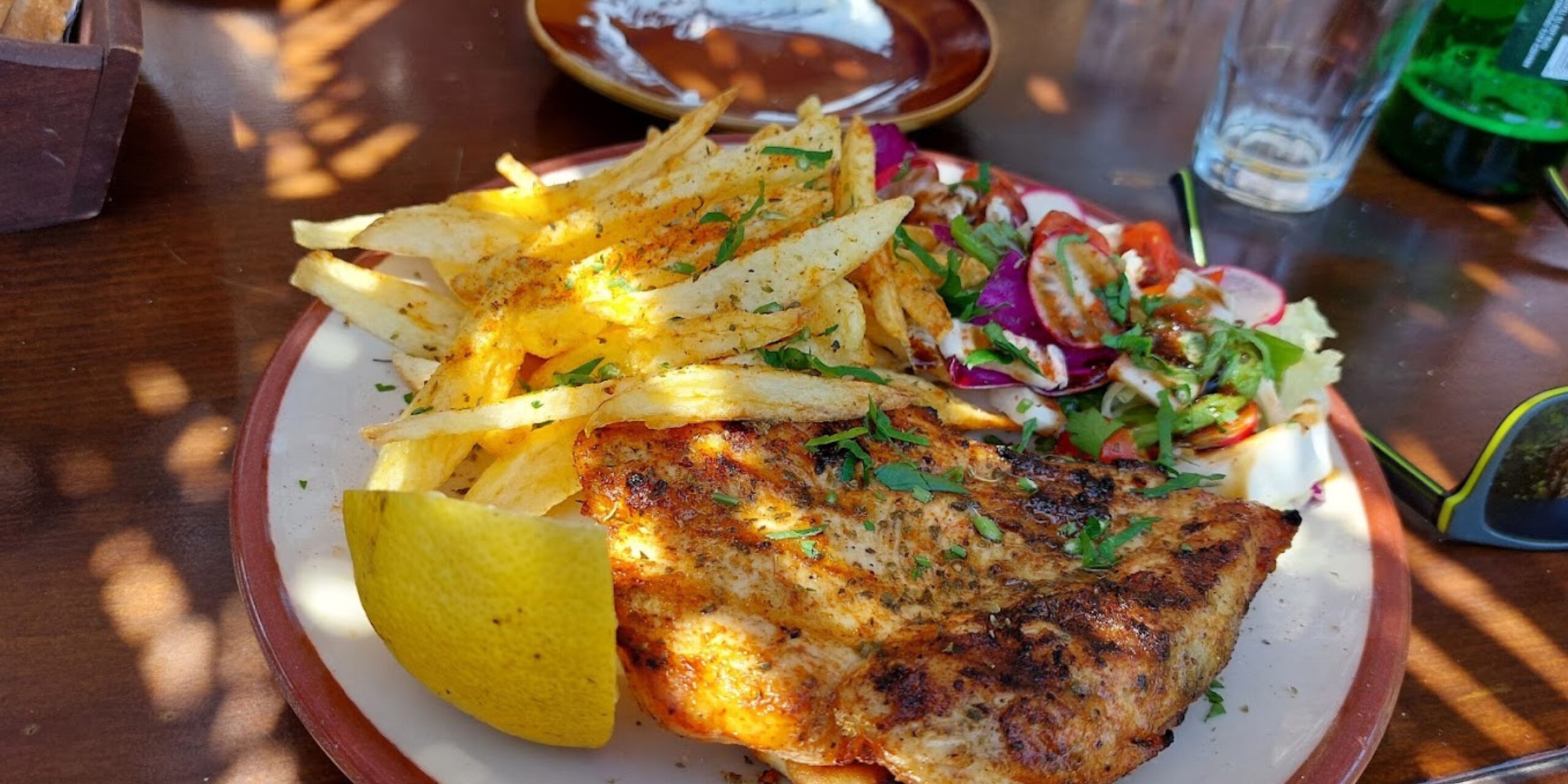Plate of grilled fish with lemon, fries, and a side salad on a wooden table.