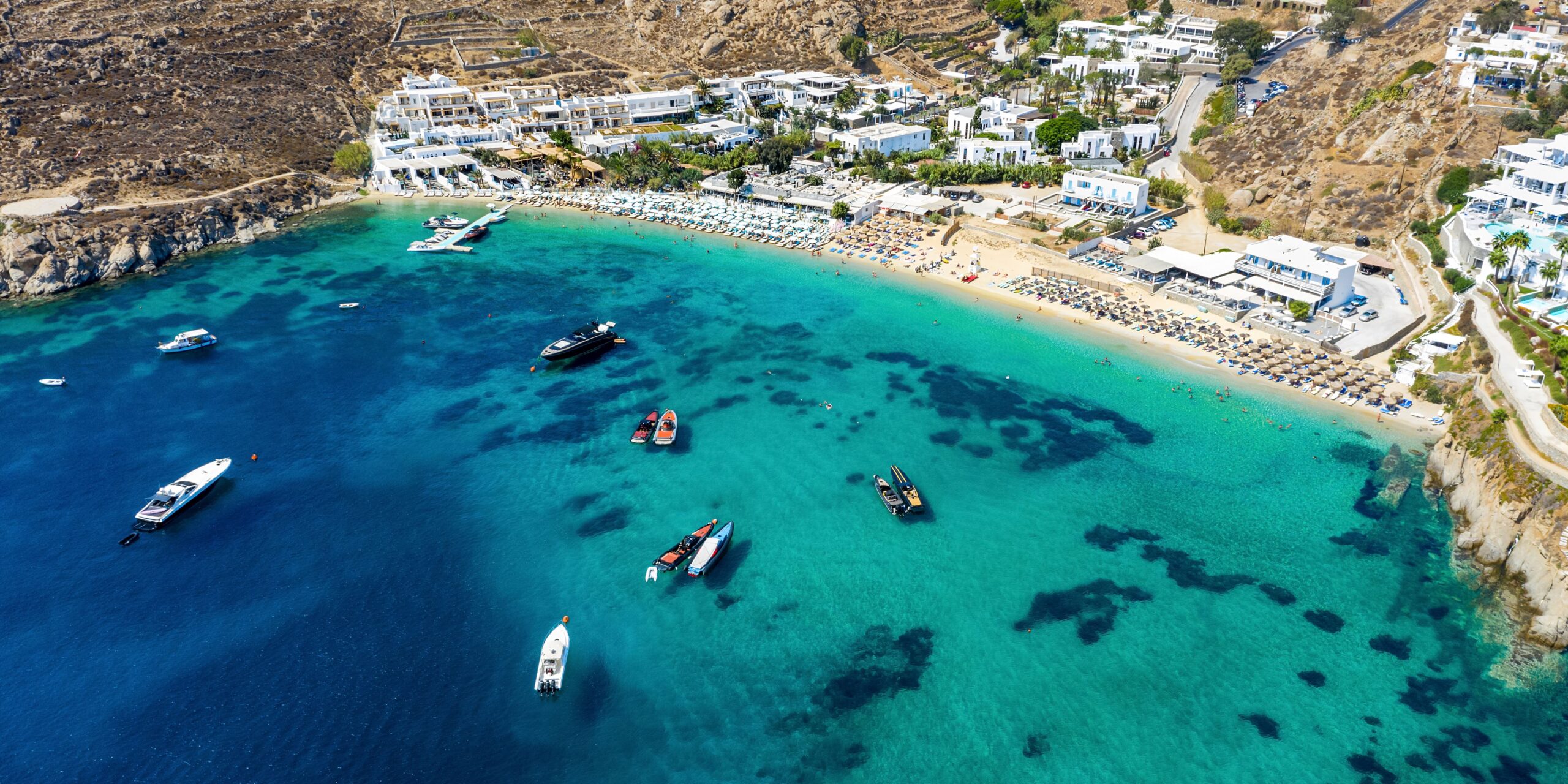 Boats float on the crystal-clear waters of a bustling Mykonos beach, a favorite spot for sun-seekers and sea-lovers.