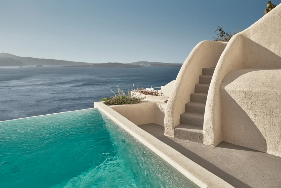 Side view of an infinity pool with stairs leading up to a traditional whitewashed structure, overlooking the sea.