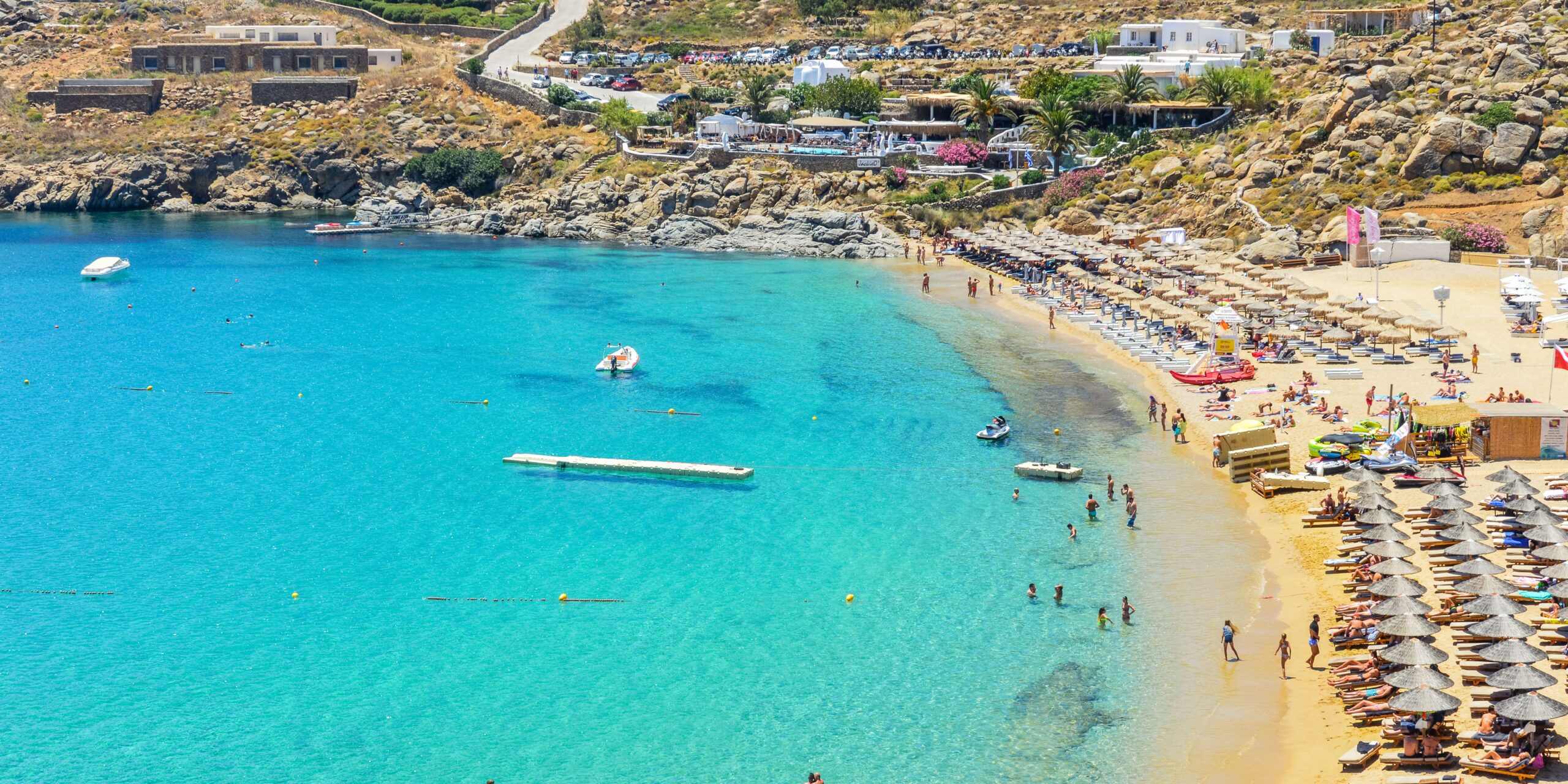 Beachgoers enjoy the sun and clear turquoise waters at the bustling Paradise Beach in Mykonos.