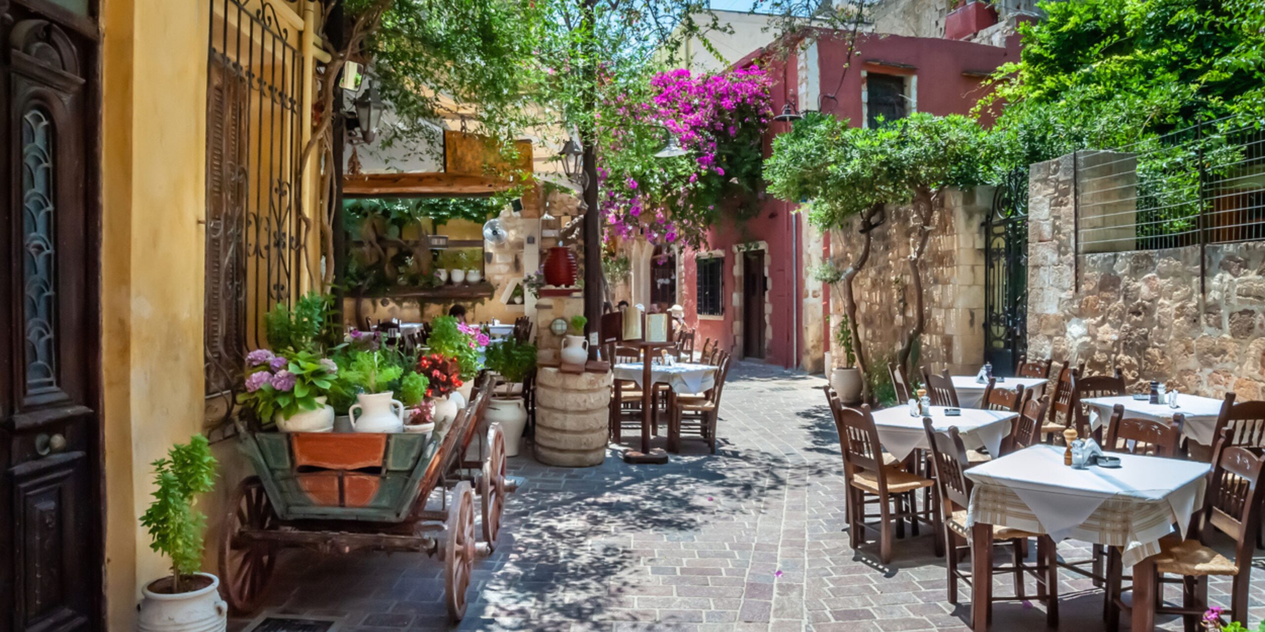 Charming alley with outdoor dining tables, plants, and blooming flowers in an old town.