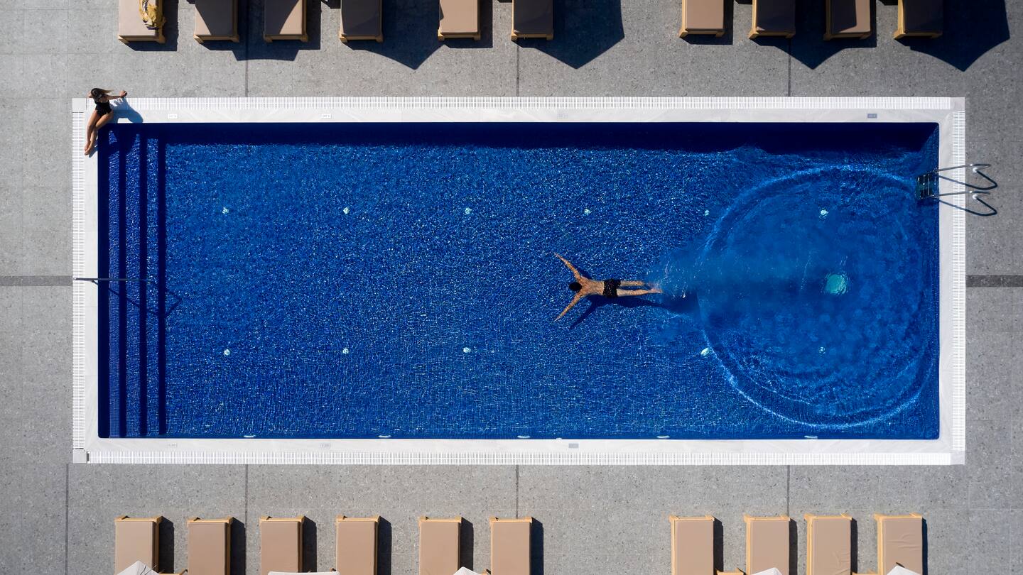 Aerial view of a person swimming in a long rectangular pool with surrounding sun loungers.