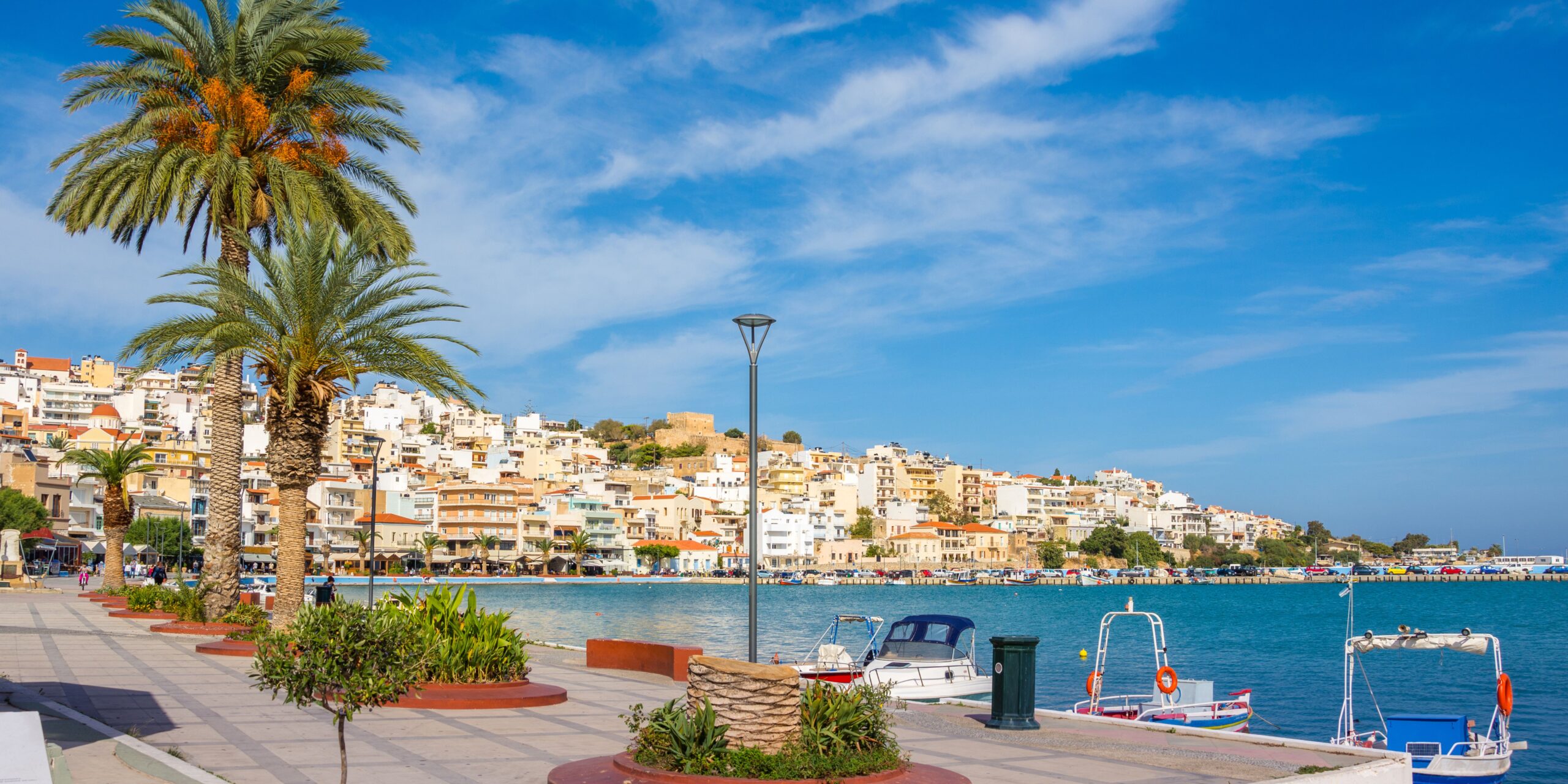A seaside promenade with palm trees, street lamps, and boats moored along the waterfront, with a view of a densely built hillside town under a blue sky.
