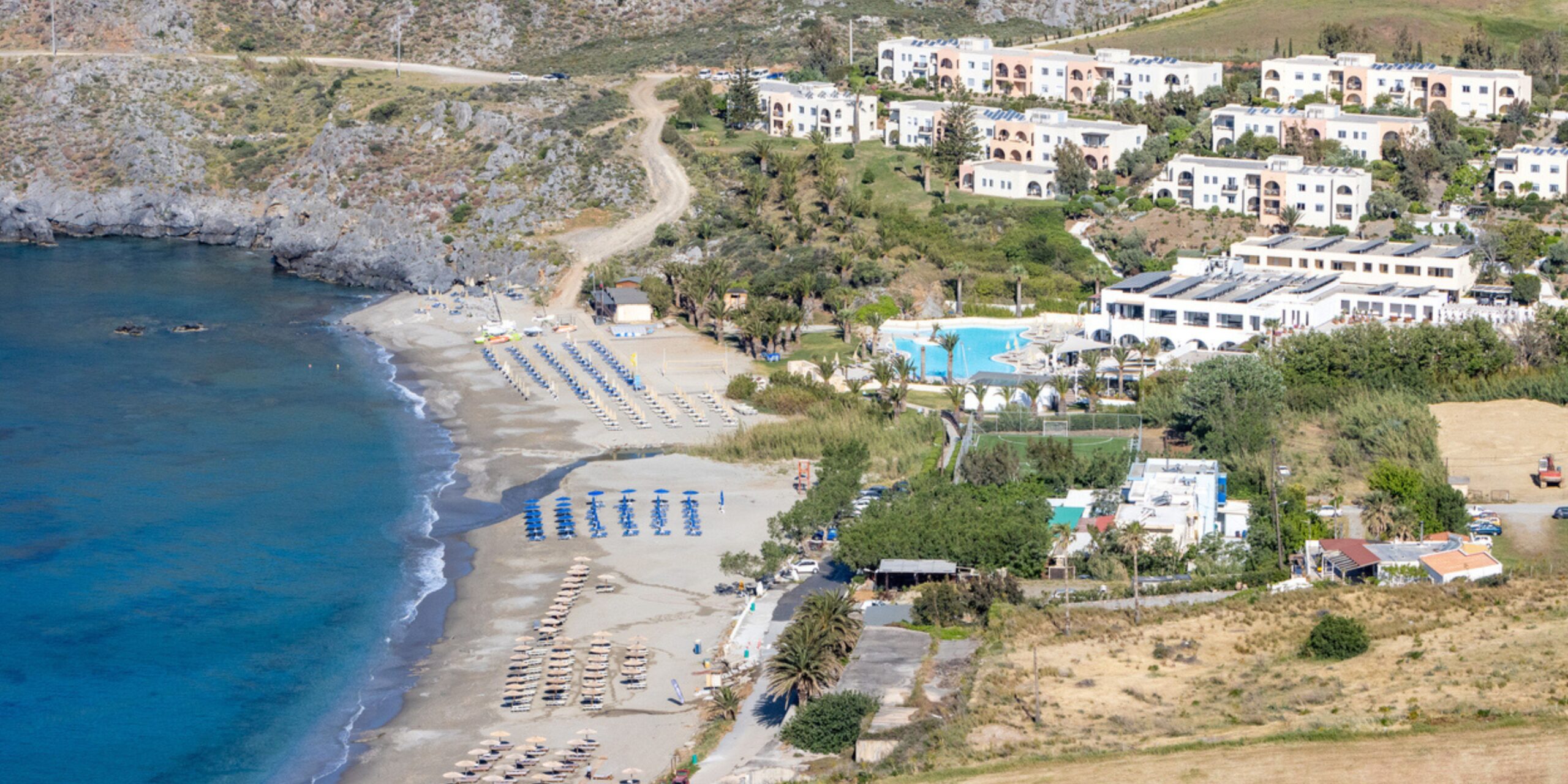 Aerial view of a coastal resort with a swimming pool, beach chairs lined up on the sand, and Mediterranean-style buildings nestled in a hilly landscape.