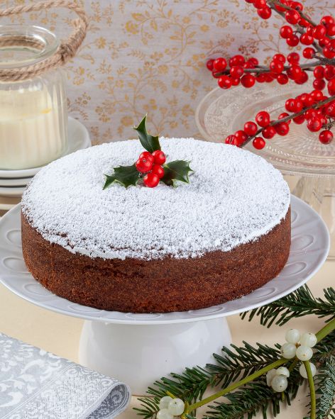 A freshly baked round cake dusted with powdered sugar sits atop a white cake stand, giving the appearance of a light snowfall. It's adorned with a sprig of holly with bright red berries, adding a festive touch. The cake stand is placed on a table with a subtly patterned golden beige tablecloth, accompanied by a jar and a lit candle, enhancing the cozy holiday ambiance. In the background, vibrant red winter berries and greenery further contribute to the festive seasonal decor.