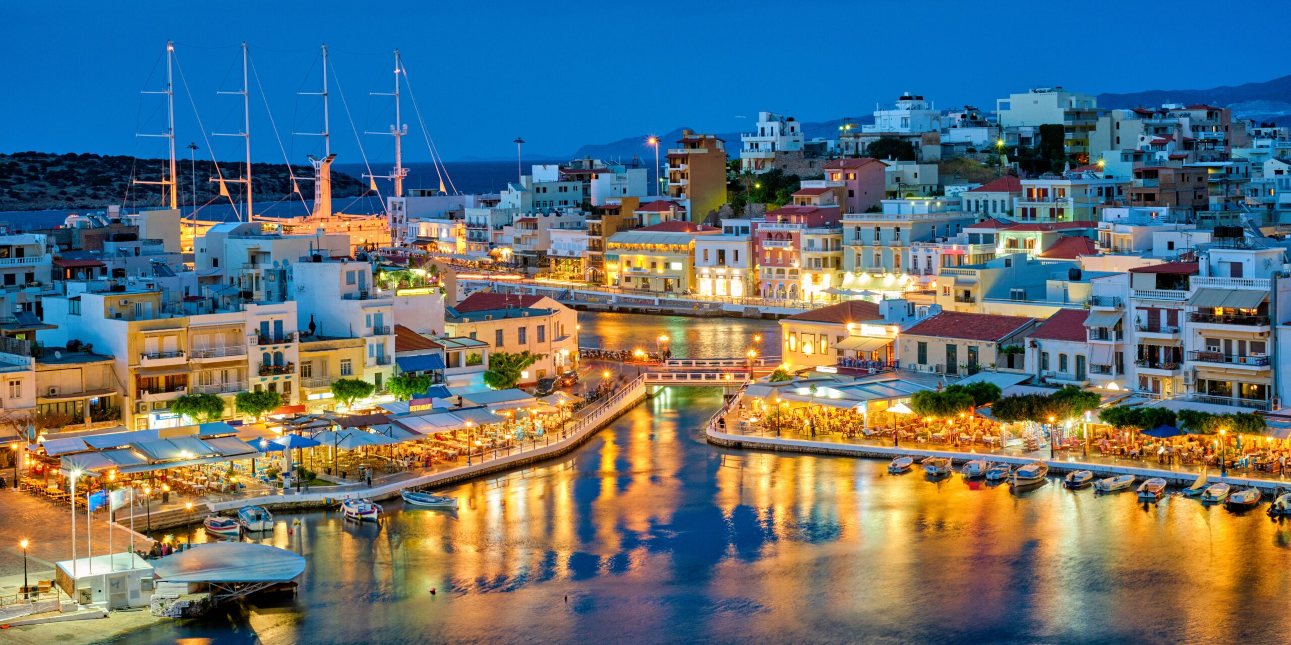 Evening view of a lit-up waterfront with buildings and moored sailboats in a Greek town.