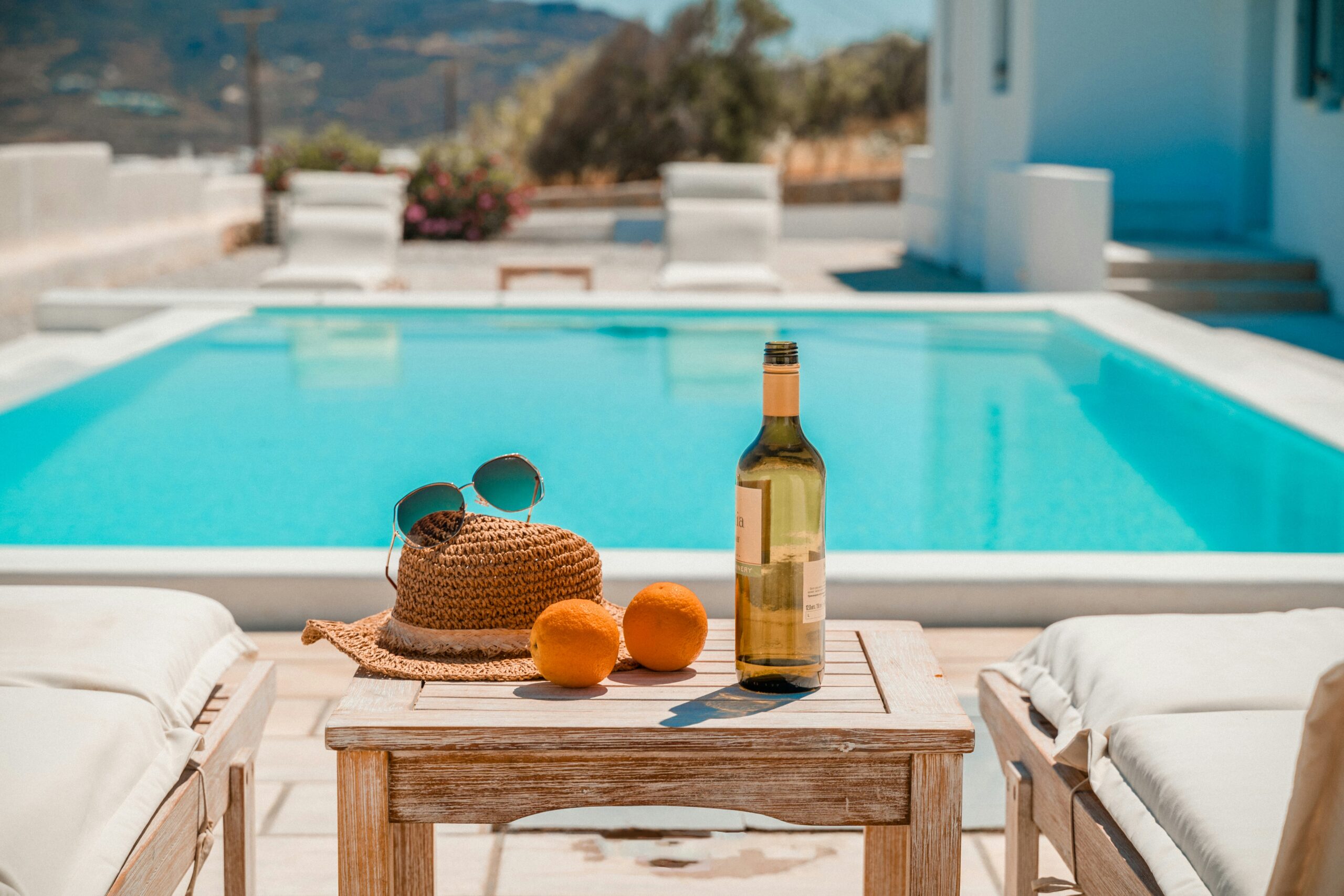 Ready for a refreshing day, a table by the poolside is set with a bottle of wine, oranges, and sunglasses, capturing the essence of a leisurely vacation.