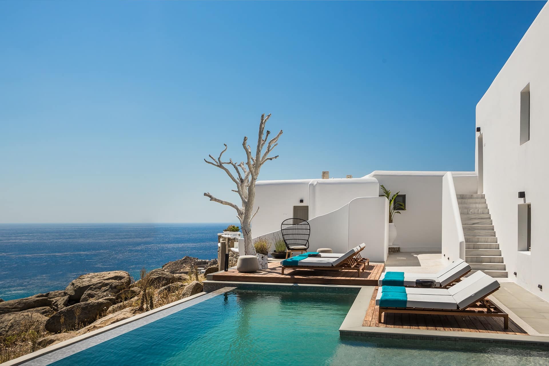 The photo shows a luxurious Mykonos hotel room decorated in white with a balcony overlooking the ocean and a swimming pool.