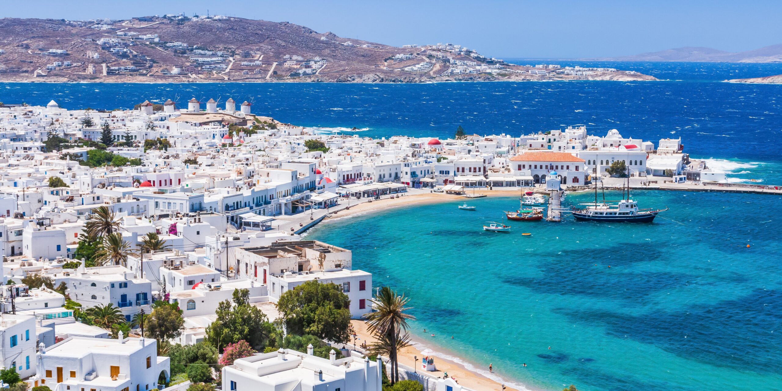 Overlooking the vibrant Mykonos town, with its white buildings clustered around a turquoise bay, where boats gently bob in the breeze.
