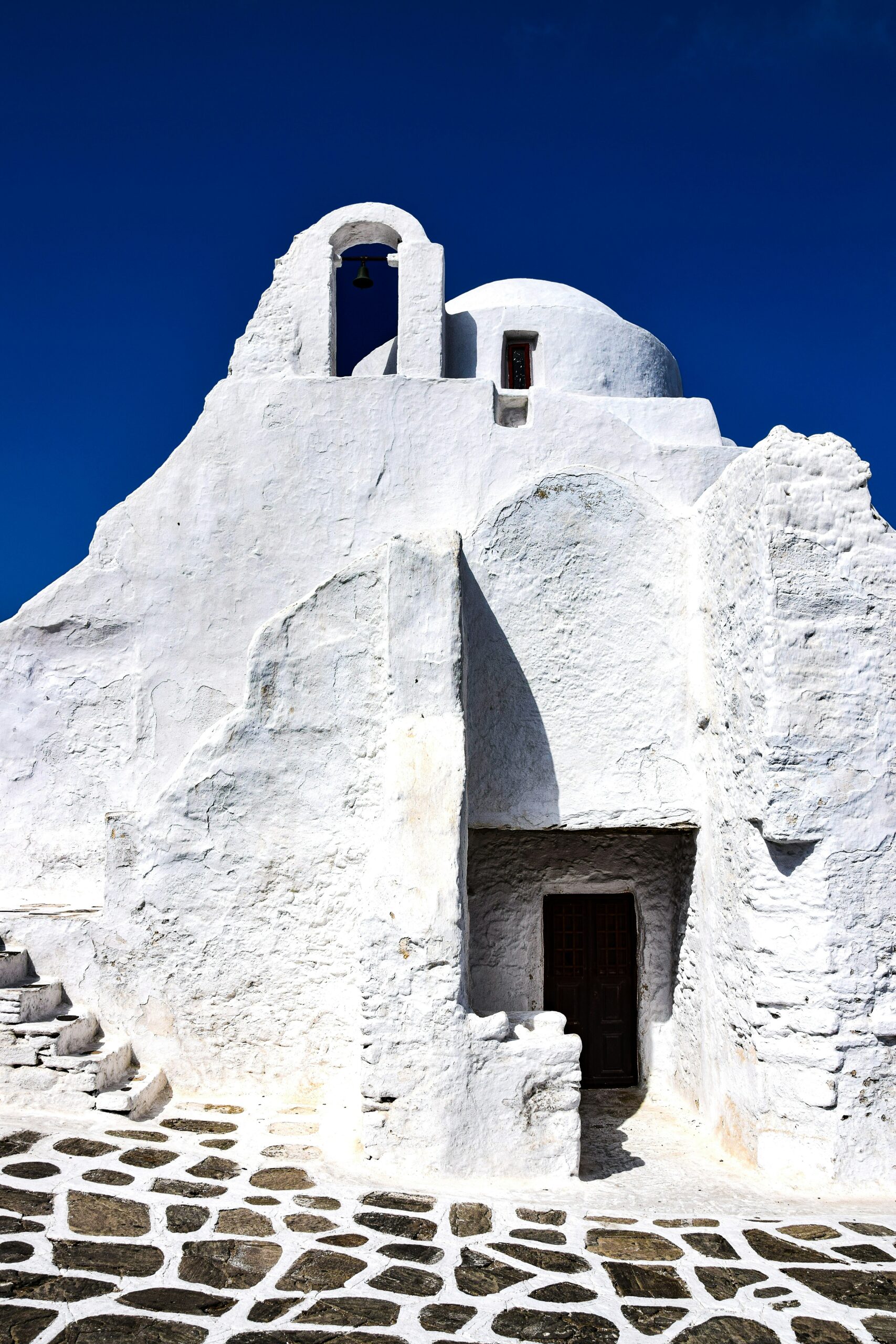 The iconic Paraportiani Church in Mykonos, Greece, stands proudly against a clear blue sky. Its striking white walls, characteristic of Cycladic architecture, glow in the sunlight. The church's smooth curves and rounded domes, painted in a dazzling white, are accentuated by the contrasting dark wooden door and window frames. In the foreground, the traditional Greek patterned cobblestone path leads up to the entrance, inviting onlookers into this historic and serene sanctuary.