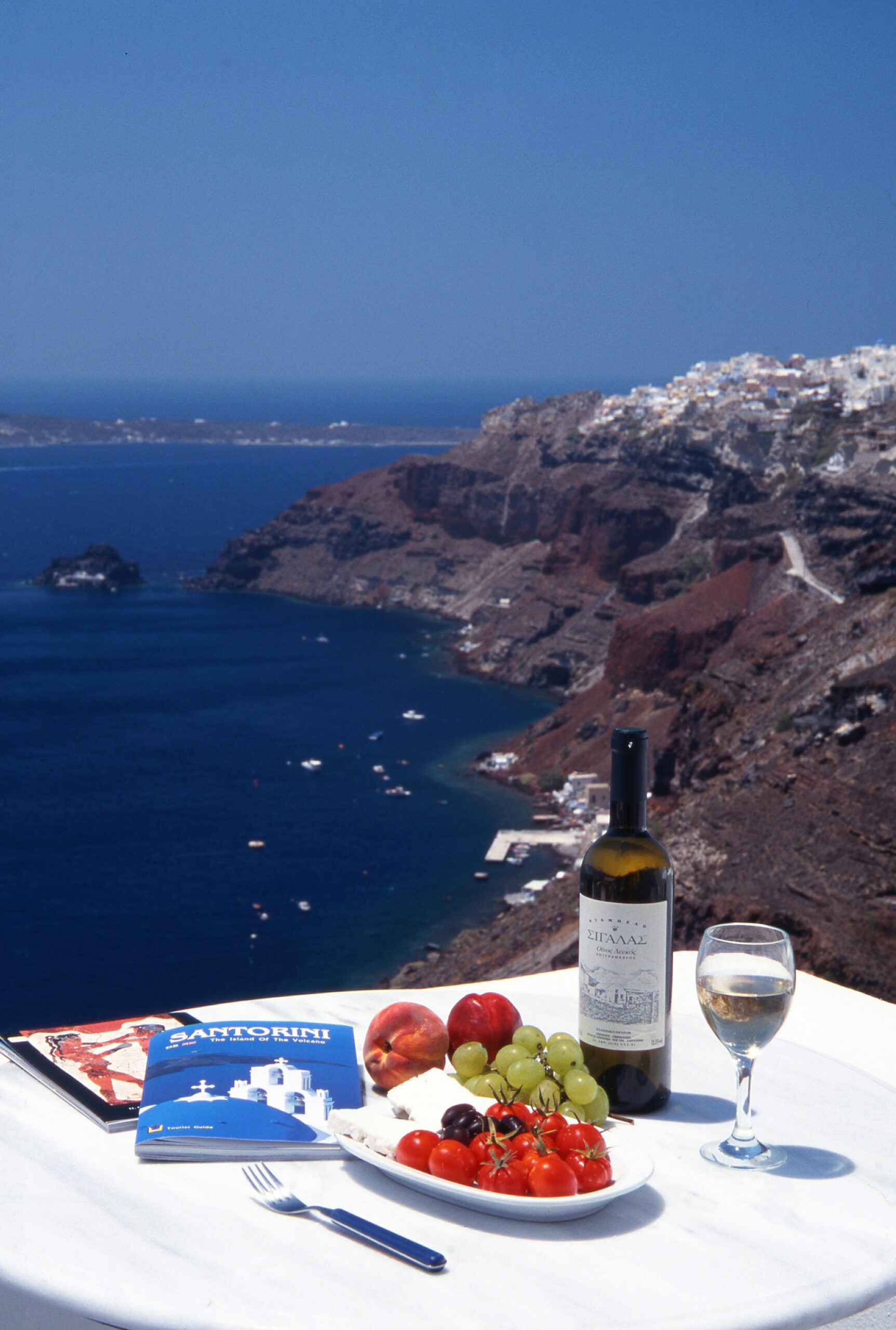 A table set with wine, fruit, and a guidebook, overlooking the cliffs and sea of Santorini.