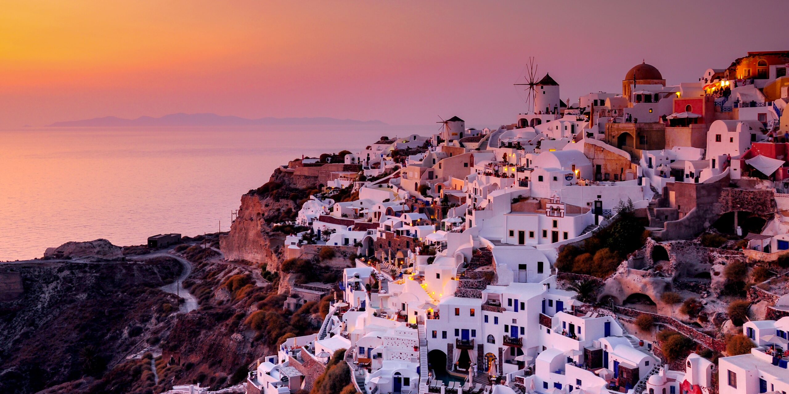 Sunset over the white buildings and windmills of Santorini's cliffside with the sea in the background.