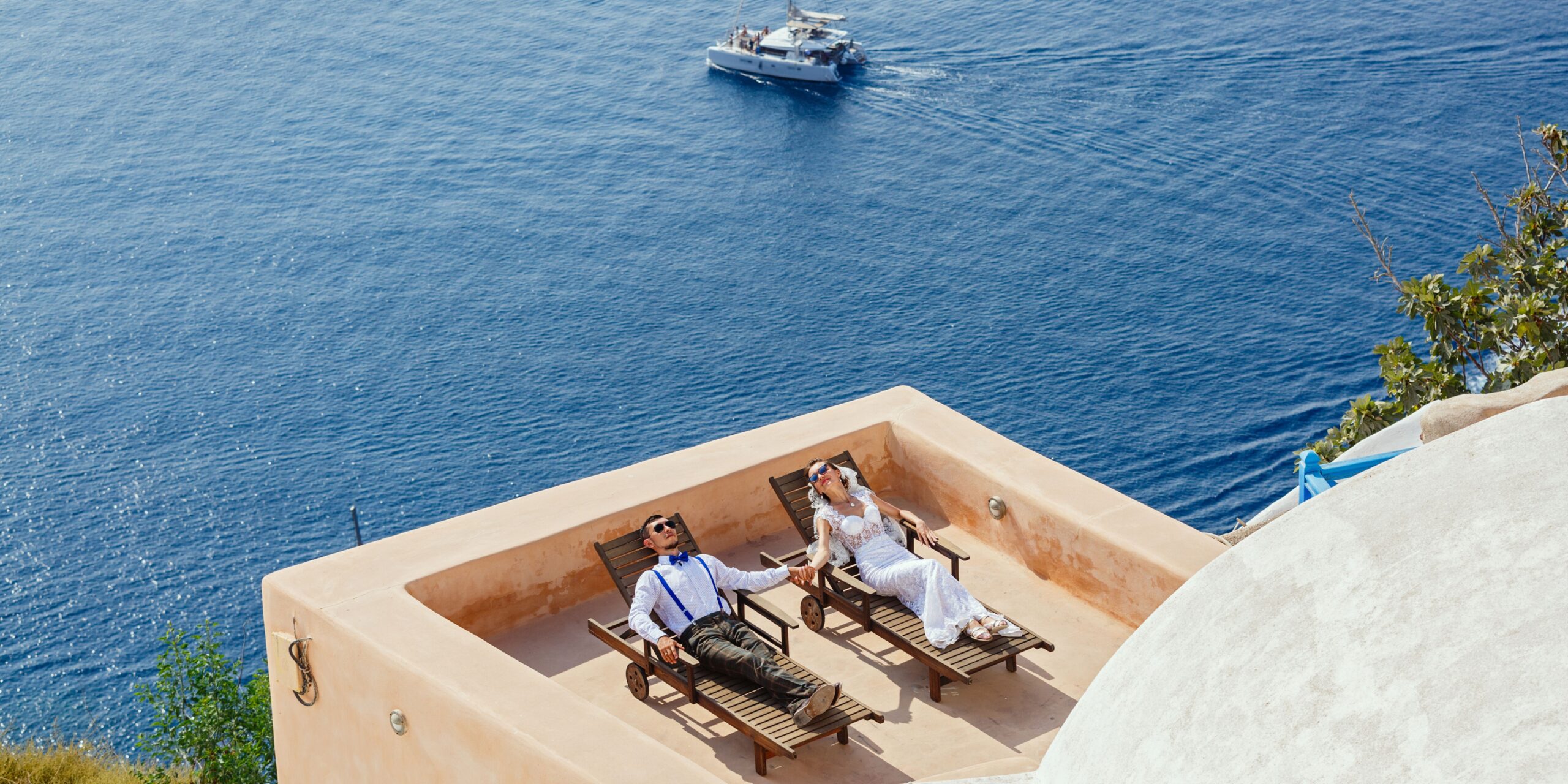 a couple dressed as a bride and groom overlooking the sea sitting on sunbeds getting their picture taken