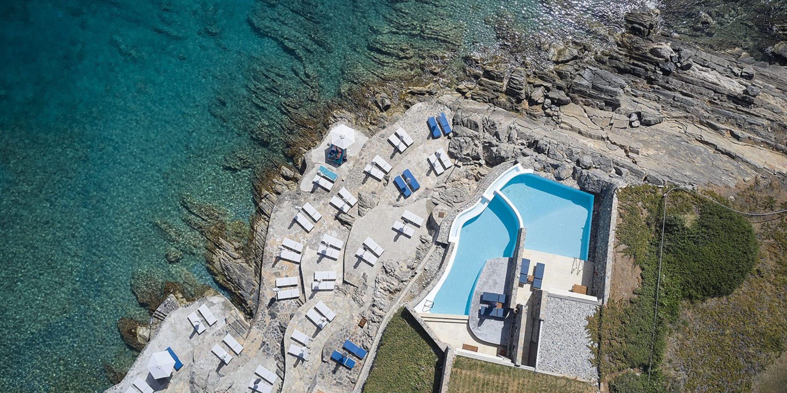 Aerial shot of a rocky shoreline with clear blue waters, featuring sun loungers and a swimming pool.