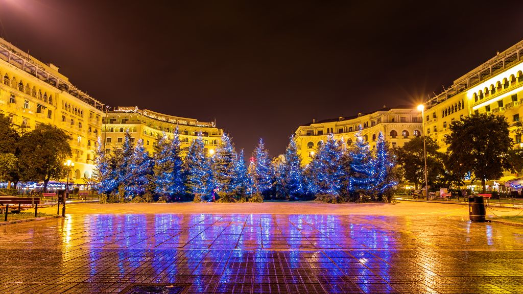 Aristotelous Square in Thessaloniki, Greece, at night, beautifully illuminated with vibrant blue lights for the holiday season. The square is wet with reflections on the ground, surrounded by grand, neoclassical buildings, creating a festive and inviting atmosphere.