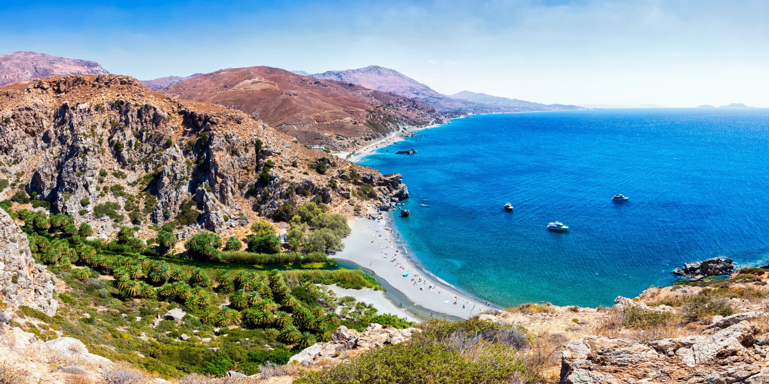 Panoramic view of a curving coastline with a blue sea, sandy beach, palm trees, and surrounding mountains.