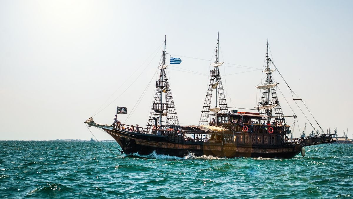 A traditional wooden sailing ship with two masts, flying the Greek flag, cruises through the blue waters off the coast of Thessaloniki, with passengers on board enjoying the sunny day.