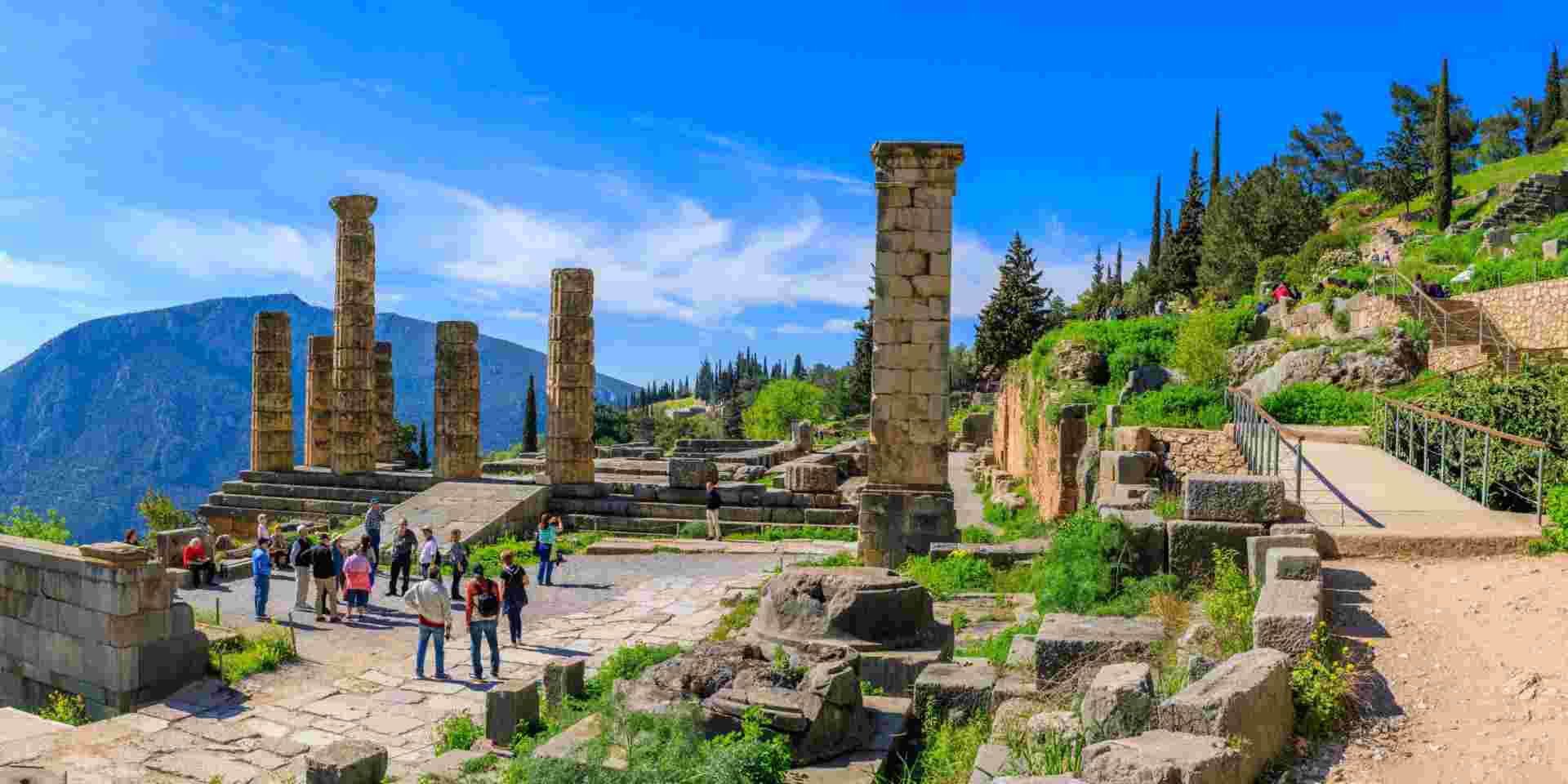 Tourists exploring the ruins of an ancient temple in Delphi, Greece, with standing columns and a mountainous backdrop under a clear blue sky. Delphi_athens to delphi day trip_feature