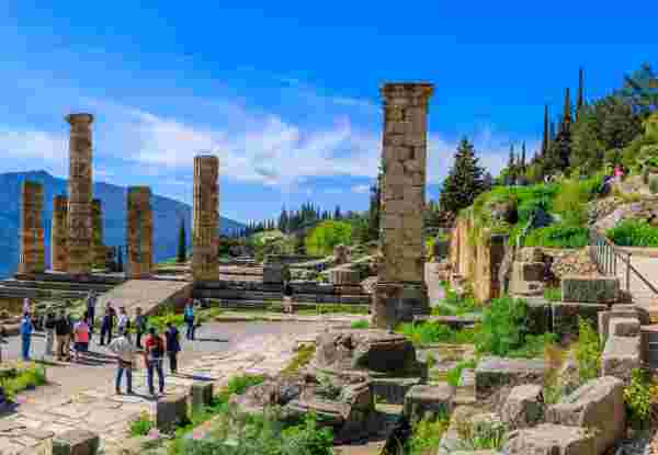 Tourists exploring the ruins of an ancient temple in Delphi, Greece, with standing columns and a mountainous backdrop under a clear blue sky. Delphi_athens to delphi day trip_feature