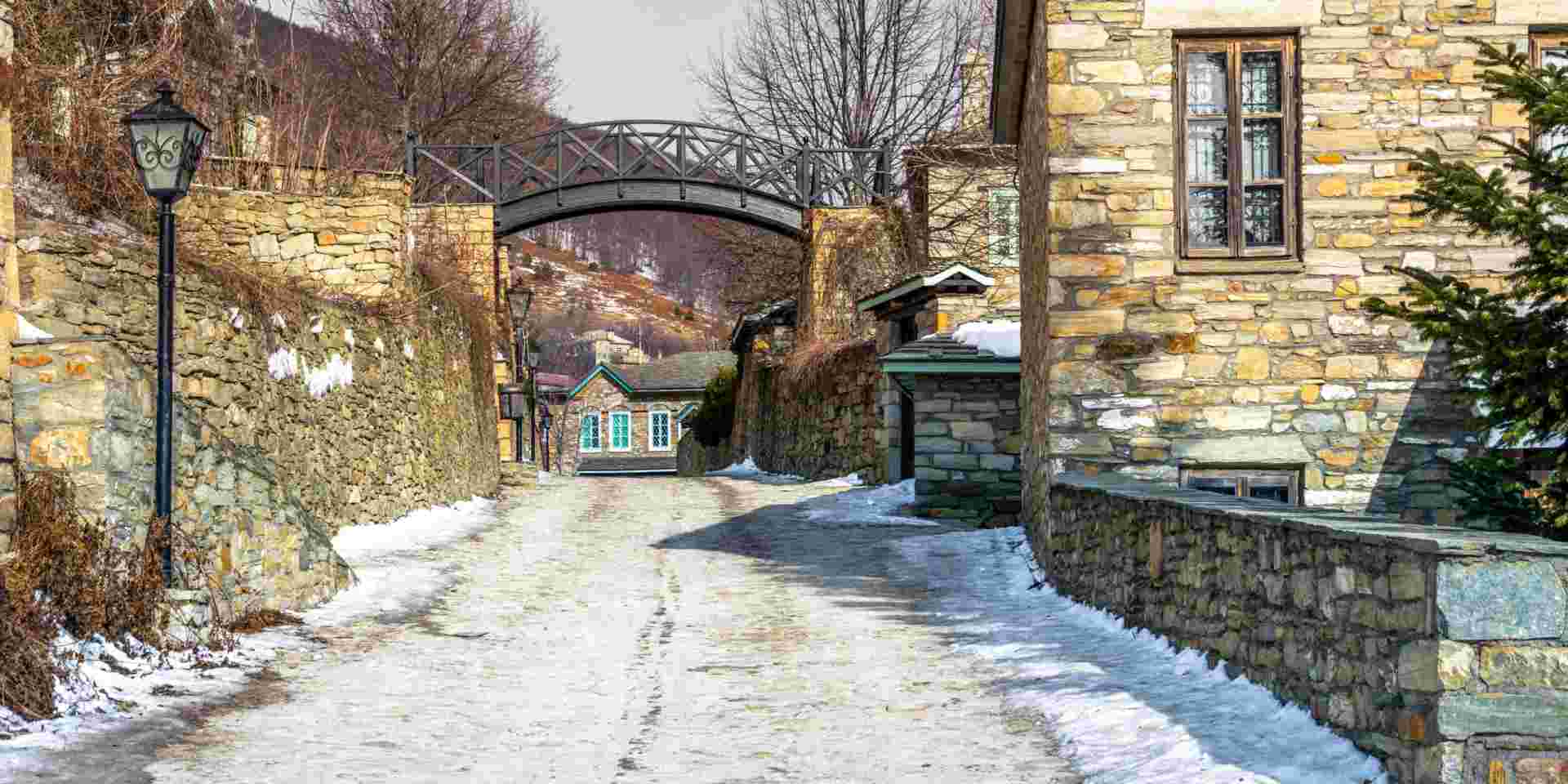 A picturesque stone-paved street in Nymfaio, lined with stone houses and walls, with a light dusting of snow, creating a peaceful winter village scene. Nymfaio village_greece in winter_feature