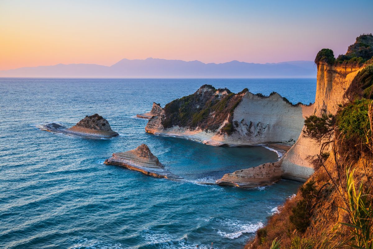 A stunning coastal view in Corfu, featuring dramatic rock formations jutting into the sea, surrounded by clear blue waters, with a colorful sunset illuminating the scene.