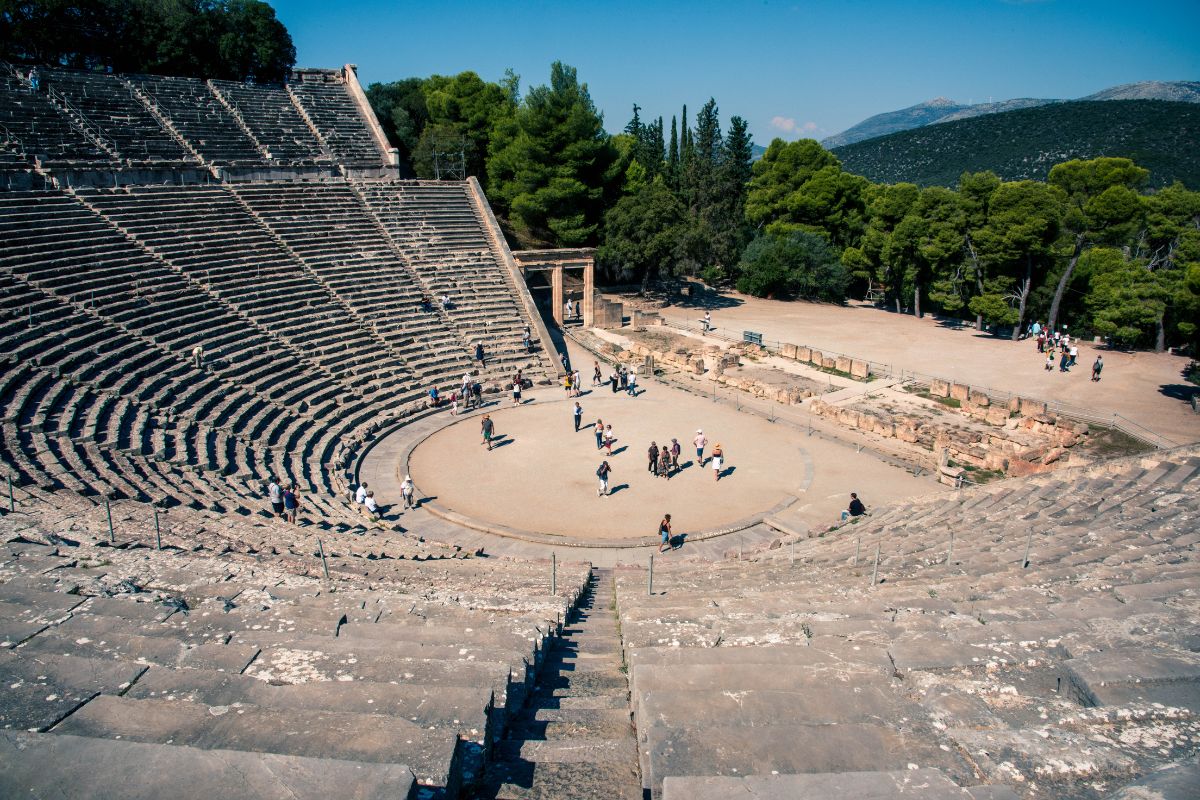 The ancient theater of Epidaurus, with its stone seating and circular stage, surrounded by lush greenery and distant hills.