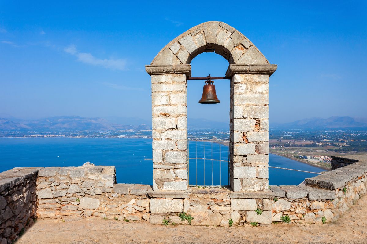 A stone arch with a bell overlooking the sea and coastline in Nafplio, Greece, under a clear blue sky.