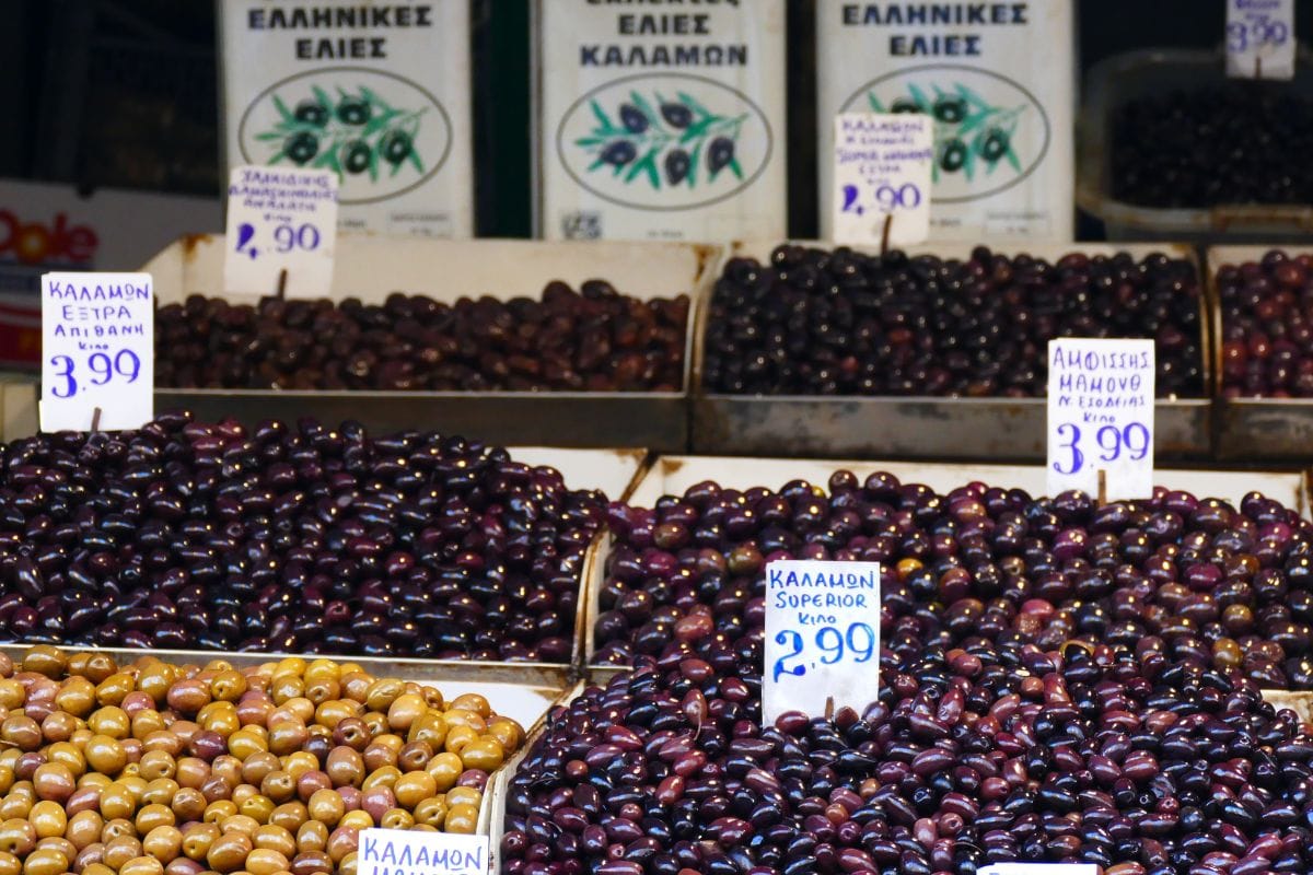 The image shows a display of various types of olives at a market in Athens. The olives are labeled with signs indicating prices and types, such as Kalamon and Amfissis, with large bins of dark and green olives on display.