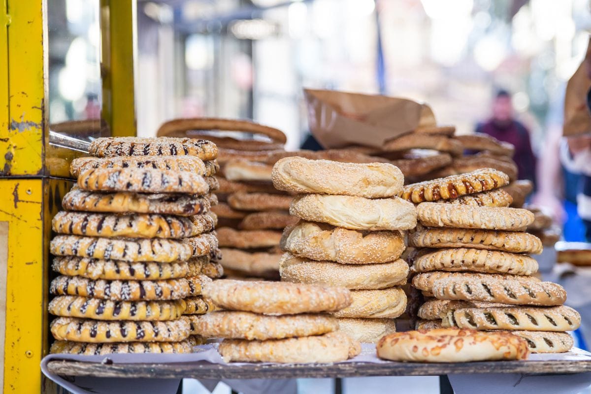 The image shows stacks of koulouri, a popular Greek sesame bread ring, displayed at a market stall in Athens. Various types of koulouri are visible, including plain and filled varieties.