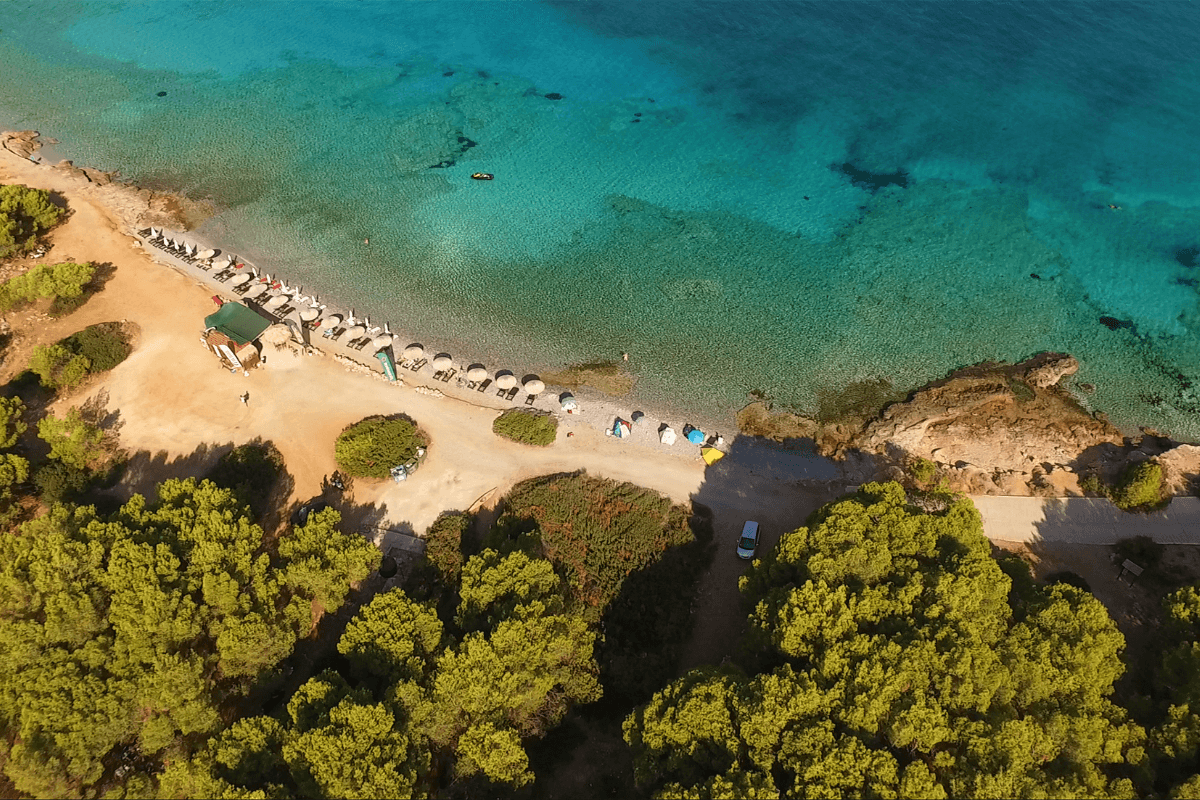 An aerial view of Agkistri Island, showcasing a clear turquoise beach lined with umbrellas and sunbeds, surrounded by lush greenery and pine trees.