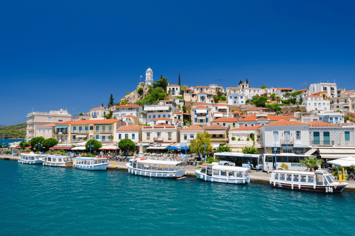 A vibrant waterfront view of Poros Island, featuring charming houses with red-tiled roofs, a hilltop clock tower, and boats docked along the harbor.