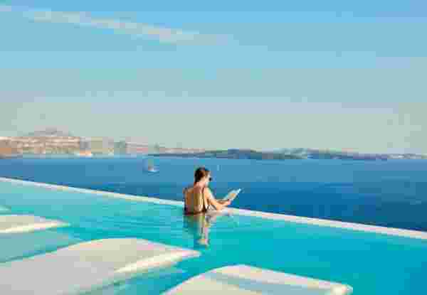 The image shows a serene infinity pool at Canaves Oia Suites in Santorini, with a person relaxing in the water while reading a book. The pool offers uninterrupted views of the caldera and the Aegean Sea, blending the luxurious setting with the natural beauty of the island. Canaves Oia Suites
