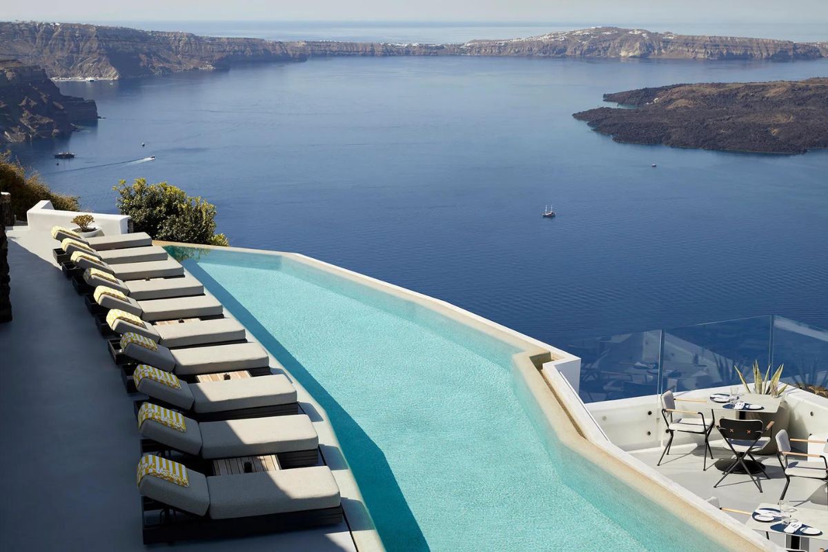 The image shows the infinity pool at Grace Hotel in Santorini, with a row of luxurious sun loungers positioned along the pool's edge. The pool overlooks the stunning Caldera and the deep blue Aegean Sea, offering a breathtaking panoramic view that epitomizes the serene beauty of Santorini.