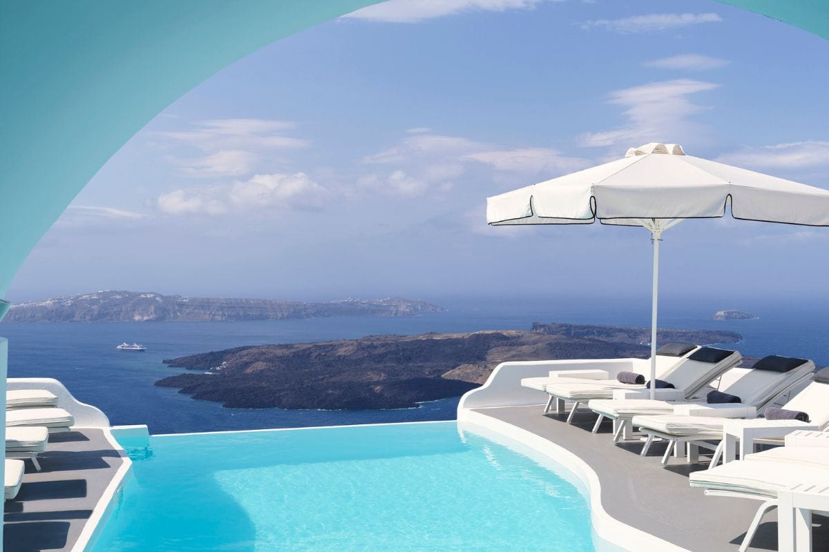 The image shows an infinity pool at Katikies Chromata Hotel in Santorini, with lounge chairs and an umbrella positioned by the poolside. The pool offers stunning views of the Aegean Sea and the caldera, framed by a soft, curved archway, creating a serene and luxurious atmosphere.