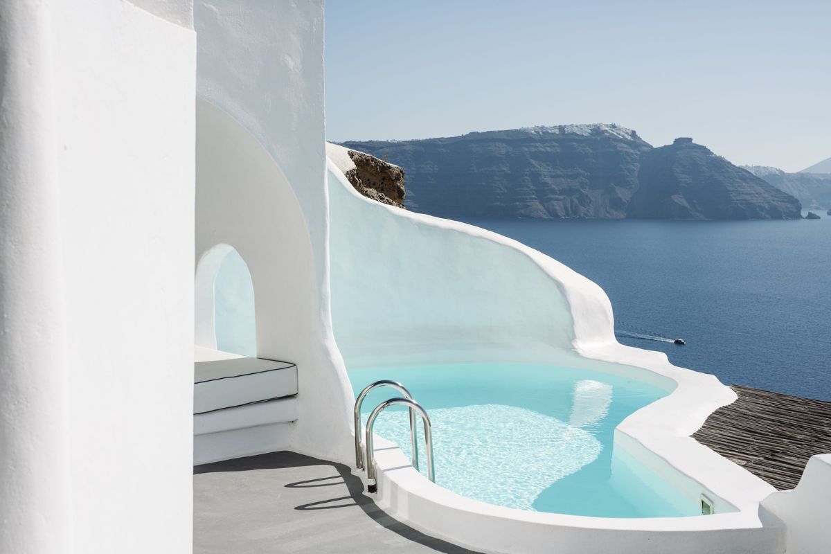 The image shows a serene, curved infinity pool at Katikies Hotel in Santorini, nestled within whitewashed, traditional architecture. The pool overlooks the stunning caldera and the Aegean Sea, creating a peaceful and luxurious retreat with breathtaking views.