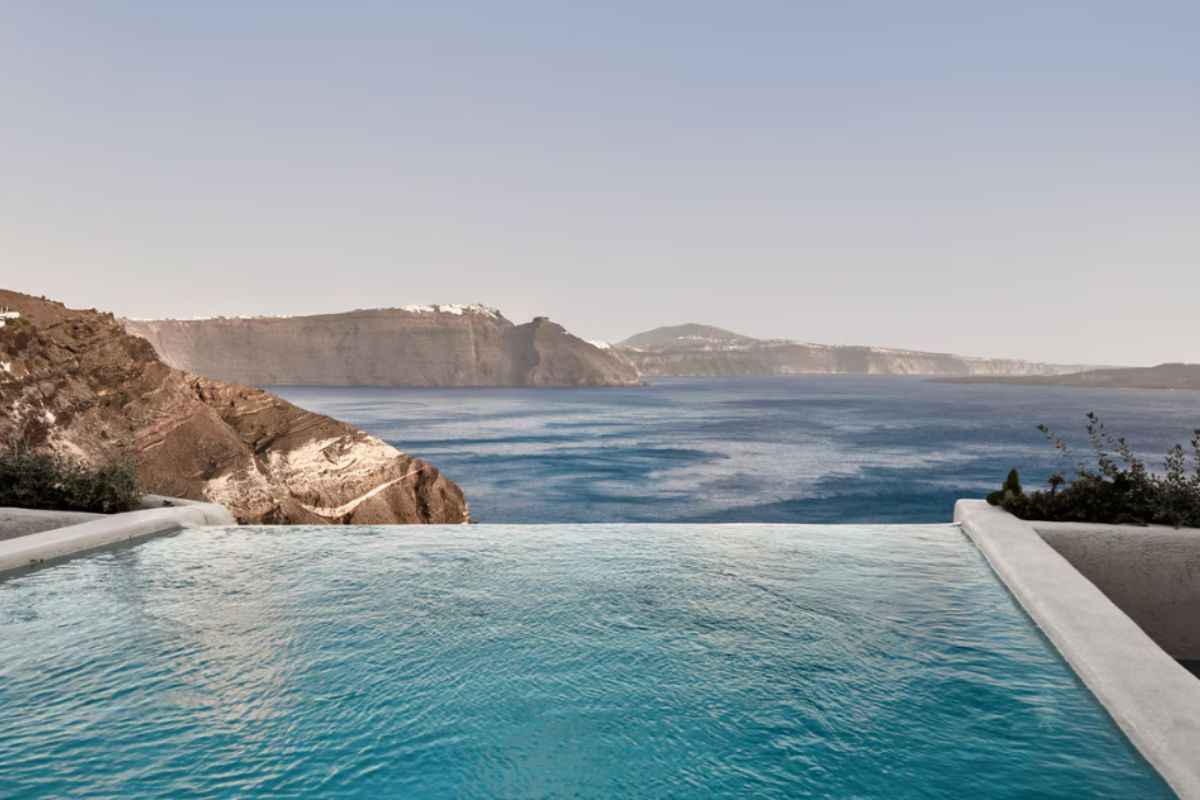 The image shows an infinity pool at Mystique Hotel in Santorini, overlooking the deep blue waters of the Aegean Sea and the dramatic cliffs of the caldera. The pool offers a tranquil and luxurious setting, seamlessly blending with the natural beauty of the surrounding landscape.