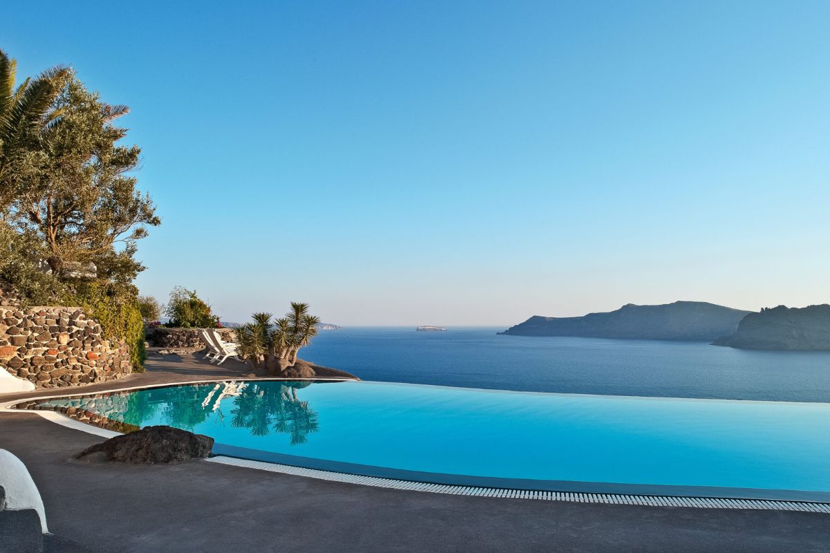 The image shows an infinity pool at Perivolas Lifestyle Houses in Santorini, offering a serene view of the Aegean Sea. The pool is surrounded by natural stone walls and lush greenery, creating a tranquil and luxurious setting with a stunning backdrop of the sea and distant islands.