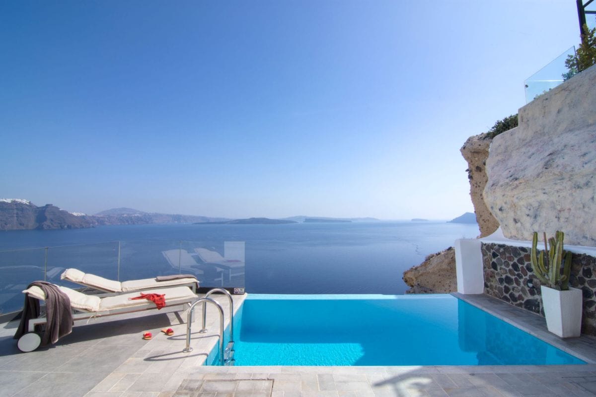 The image shows a private pool at Santorini Secret and Spa Hotel, positioned on a terrace overlooking the expansive Aegean Sea. The pool area includes a comfortable lounger and is bordered by natural rock formations, offering a luxurious and tranquil retreat with breathtaking views of the caldera and surrounding islands.