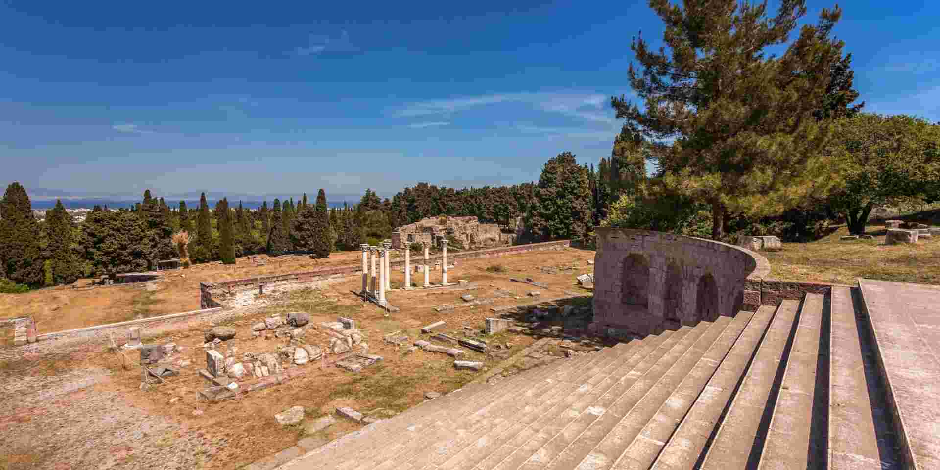 Ancient ruins on the island of Kos, featuring stone columns and scattered remnants of structures, with tall trees and a clear blue sky in the background. kos island_Best Greek Island to Visit in May_feature